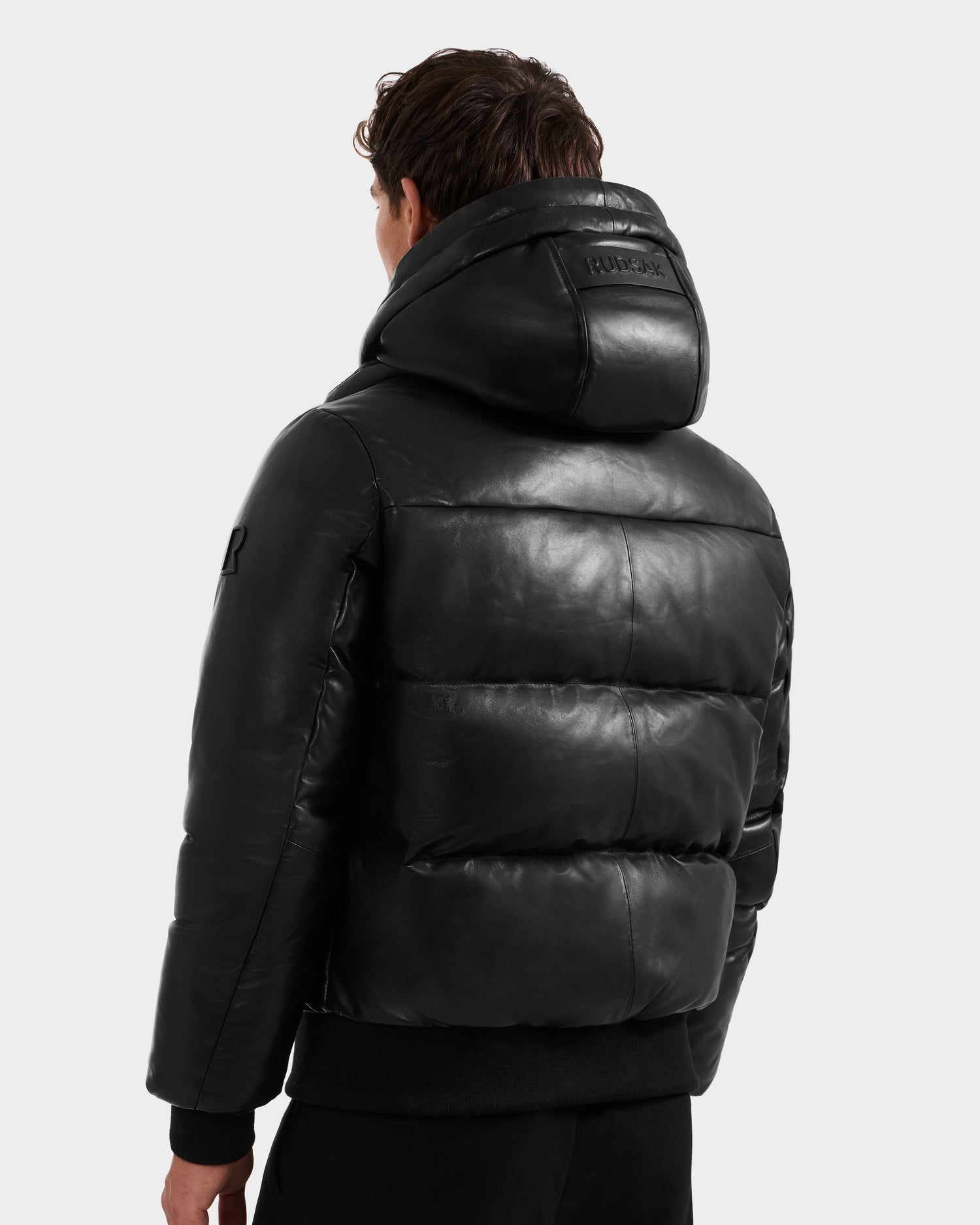A person with short brown hair is seen from the back wearing the Rudsak JORDY X - 6125694 Black leather bomber jacket, featuring a hood with a logo patch and a shiny leather-like texture against a light gray background.