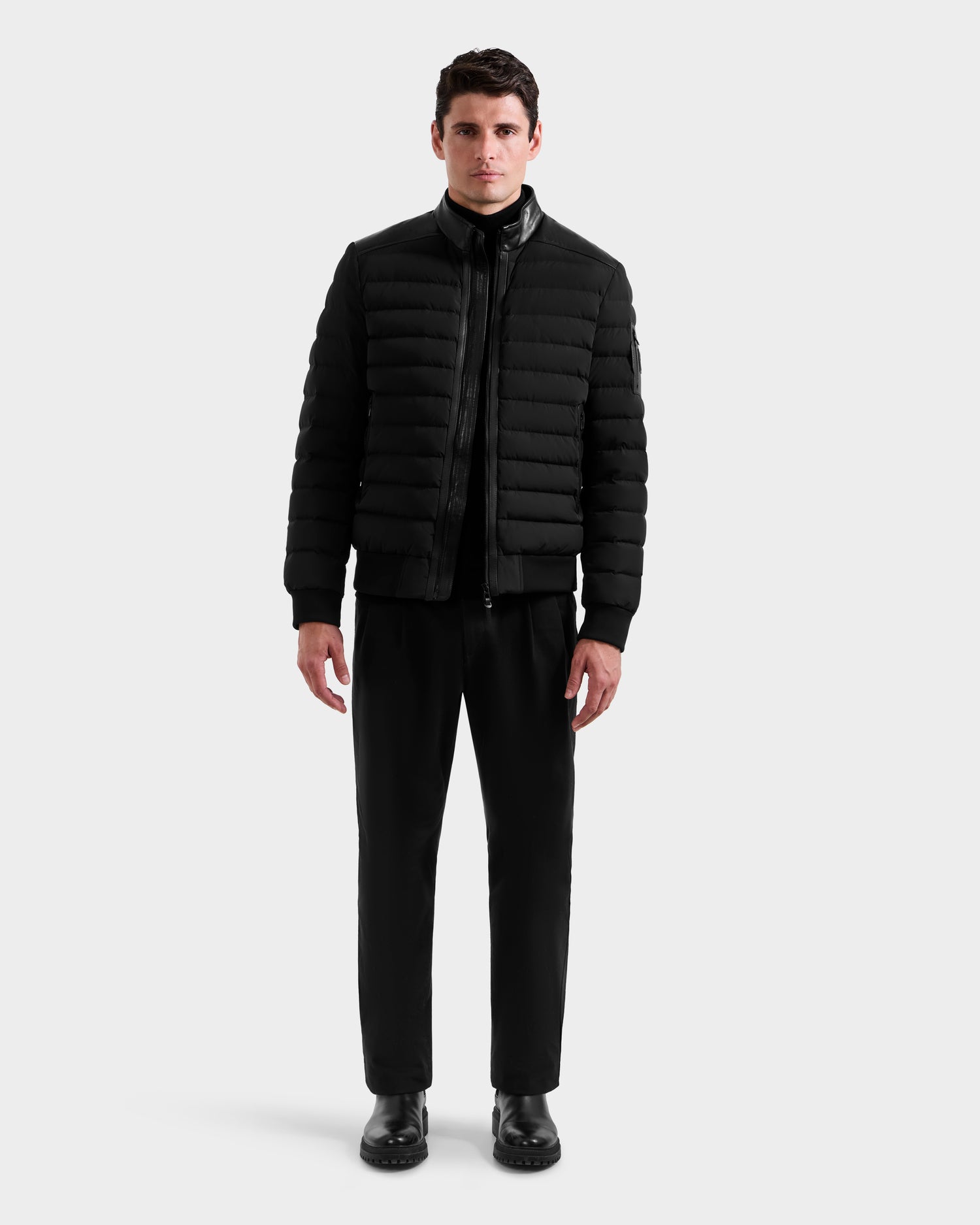 A man stands facing forward against a plain light gray background, wearing the Rudsak JOSHUA - 6125722 Black quilted down jacket, black pants, and black shoes.