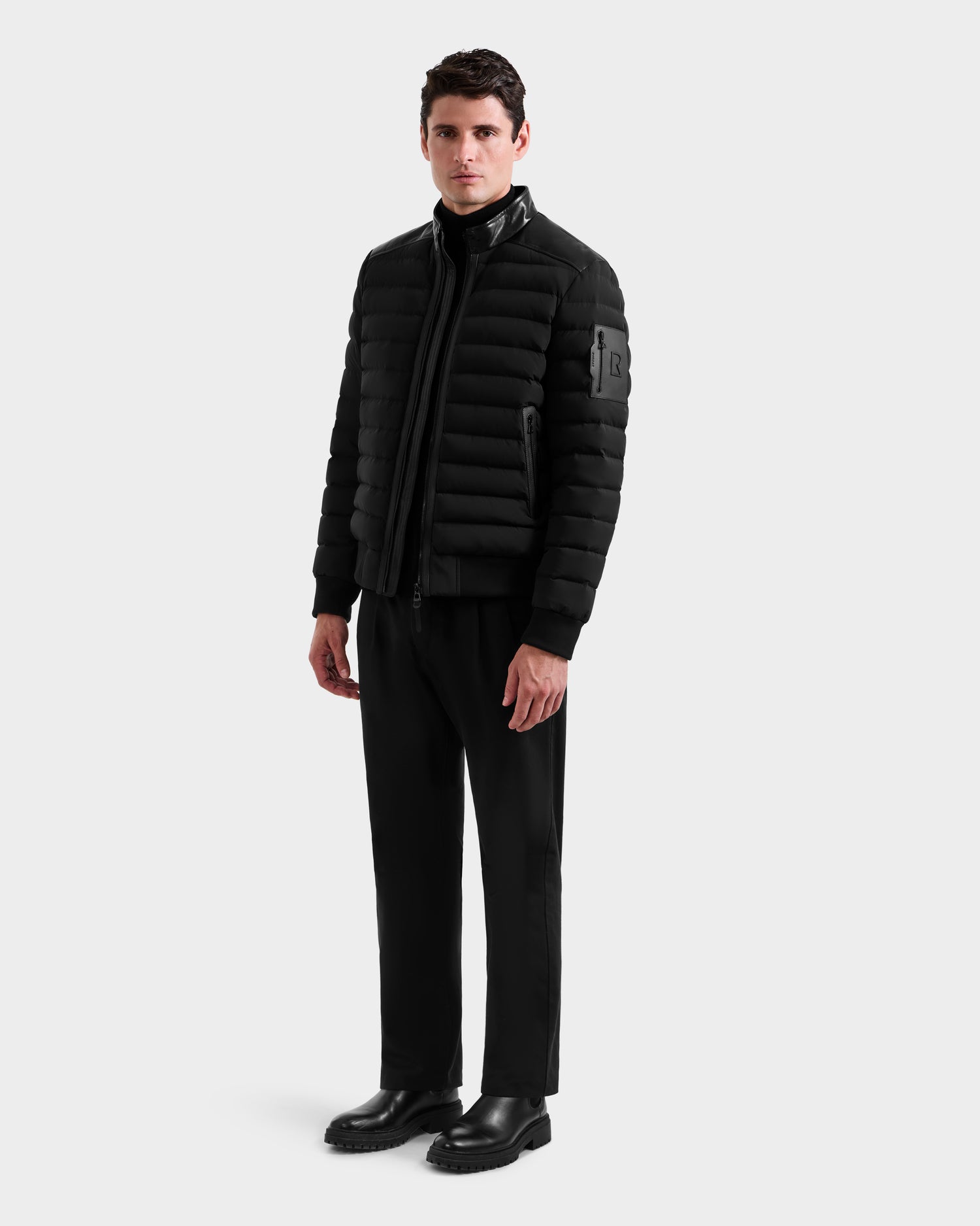 A man stands against a plain white background wearing the Rudsak JOSHUA - 6125722 Black quilted down jacket, paired with a black turtleneck, trousers, and shoes. He faces forward with arms relaxed at his sides.