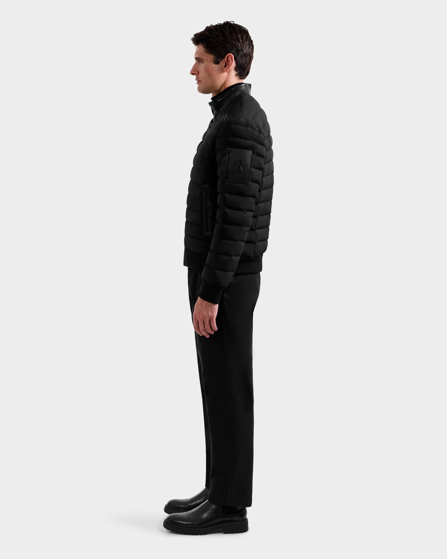 A man stands in profile facing left, wearing the Rudsak JOSHUA - 6125722 Black quilted down jacket, black pants, and black boots against a plain light gray background.