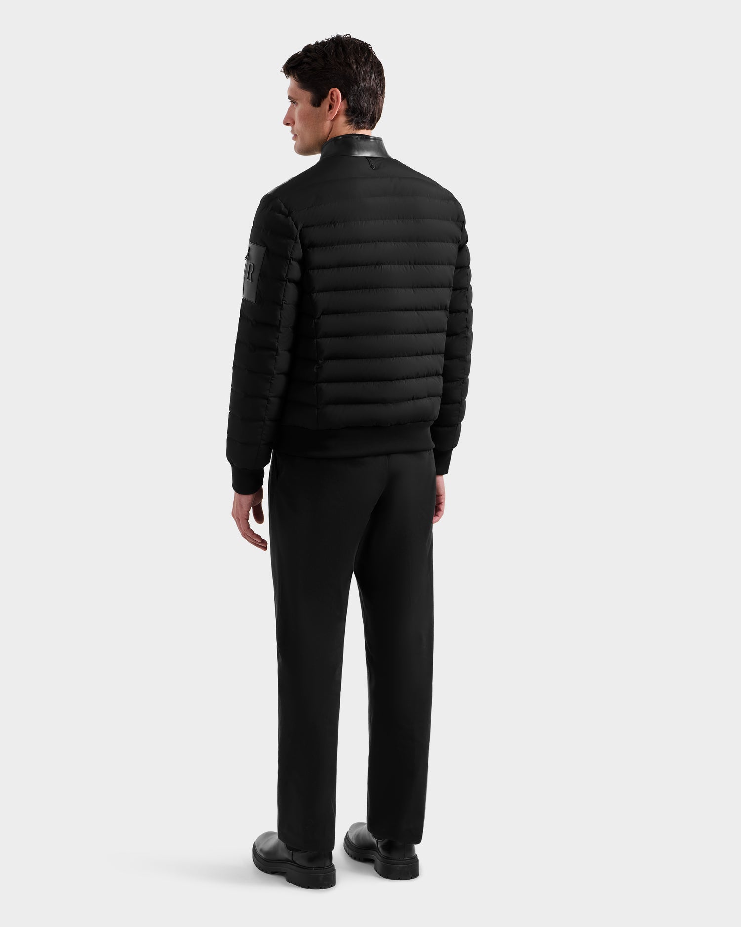 A man faces away wearing the Rudsak JOSHUA - 6125722 Black quilted down jacket with horizontal quilting and a sleeve patch, paired with black pants and shoes. The background is plain light gray.