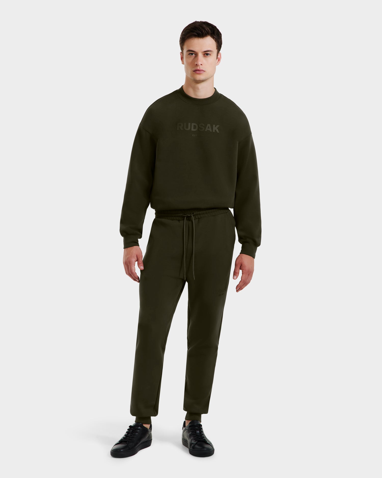 A man in the Rudsak JOSS - 9524009 Dark Olive M sweatshirt and matching joggers stands against a plain background, wearing black sneakers and gazing ahead with a neutral expression.
