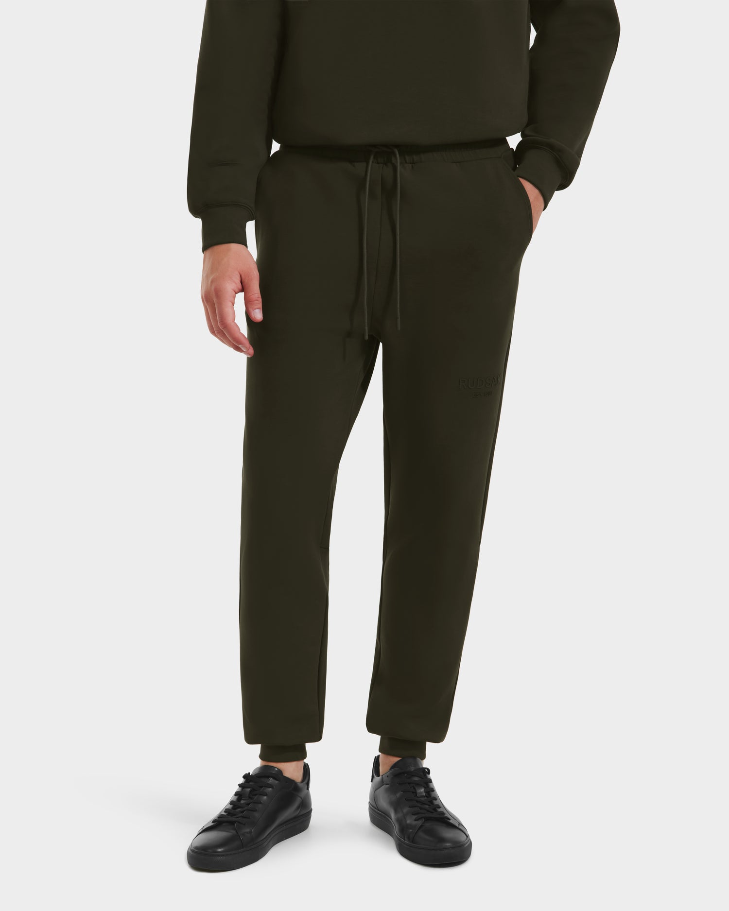 A person showcases the lower body wearing Rudsak’s JOSS - 9524009 joggers in Dark Olive M, paired with a black sweatshirt and sneakers, against a plain white background.