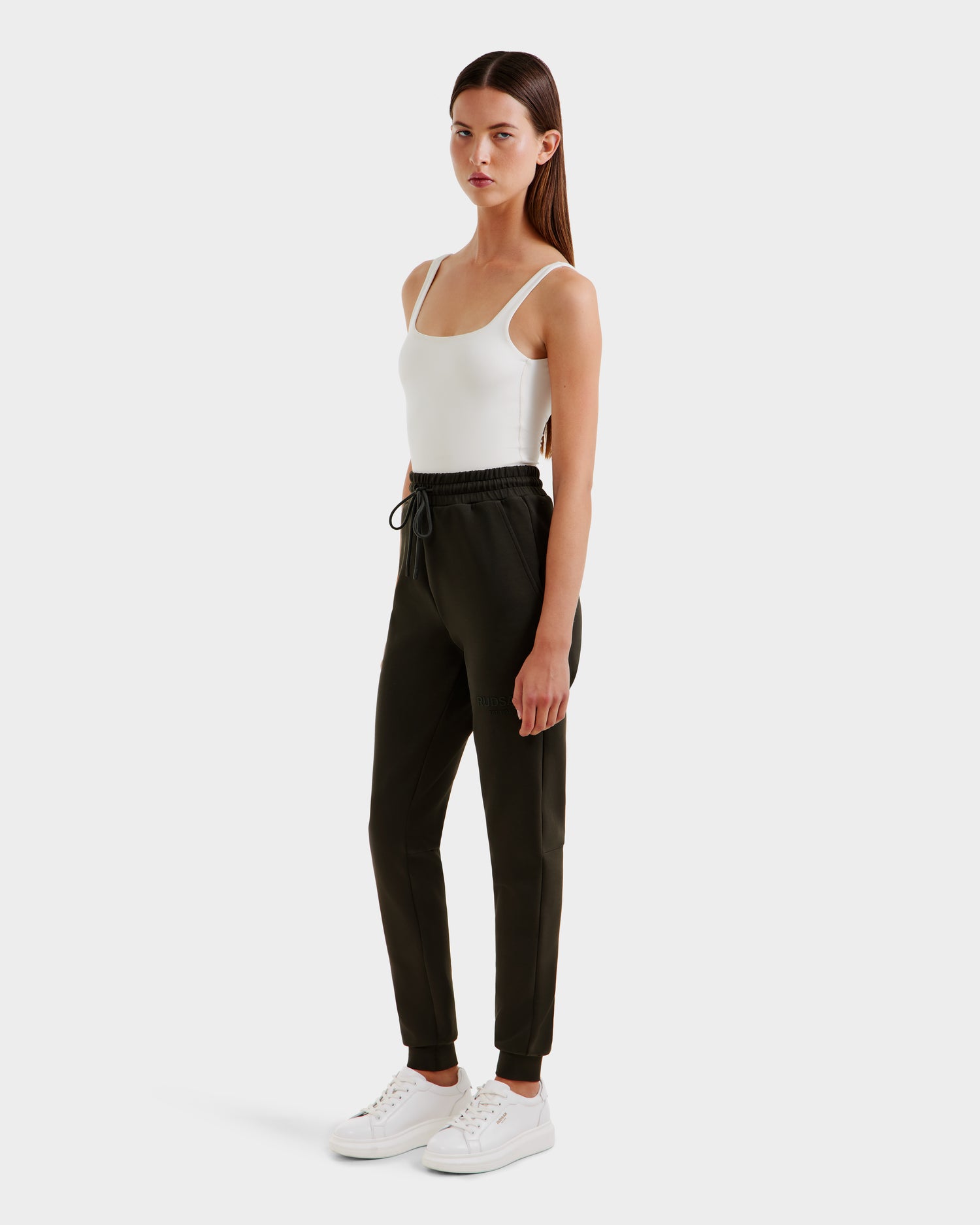 A woman stands against a plain background, wearing Rudsak’s JOSS - 9524009 Dark Olive W jogger pants with a drawstring waist, paired with a white sleeveless bodysuit and white sneakers. She faces slightly to the side with a neutral expression.