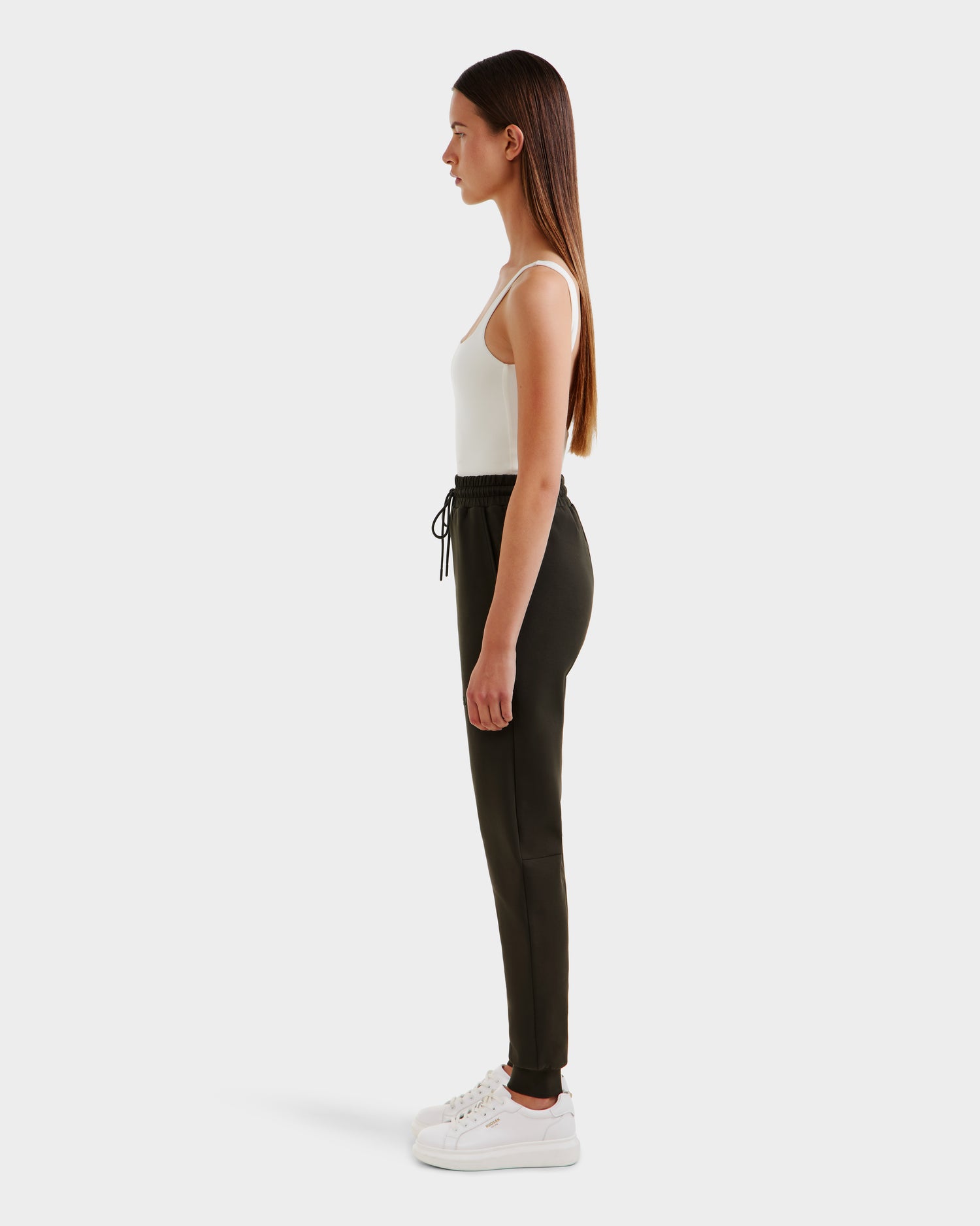 A woman with long straight hair stands in profile, wearing the Rudsak JOSS - 9524009 Dark Olive W jogger pants and a white tank top, paired with white sneakers against a plain white background.
