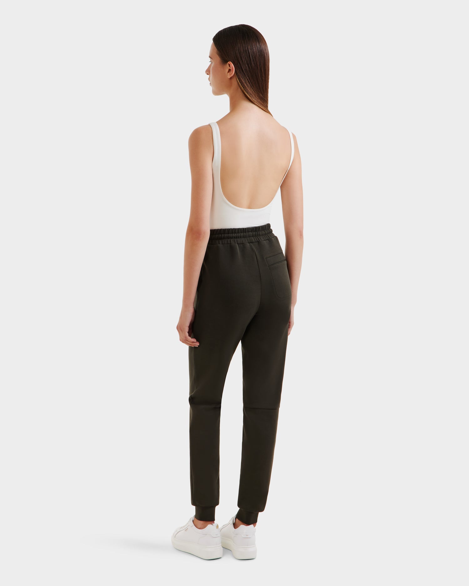 A woman faces away, wearing Rudsaks JOSS - 9524009 Dark Olive W joggers, paired with a white sleeveless low-back bodysuit and white sneakers, set against a plain light background.