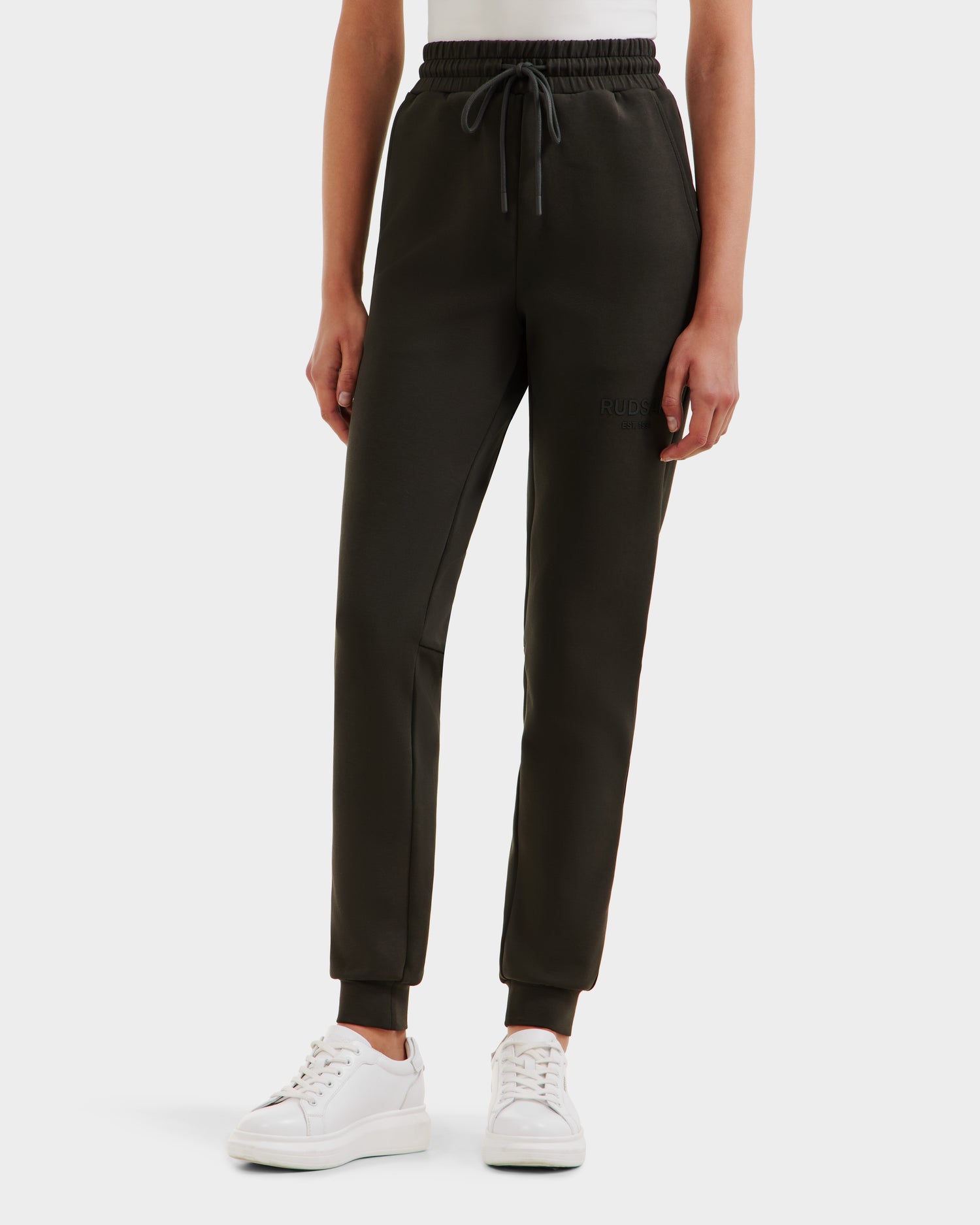 A person wearing Rudsak JOSS - 9524009 Dark Olive W joggers with a drawstring waist and white sneakers, standing against a plain white background.