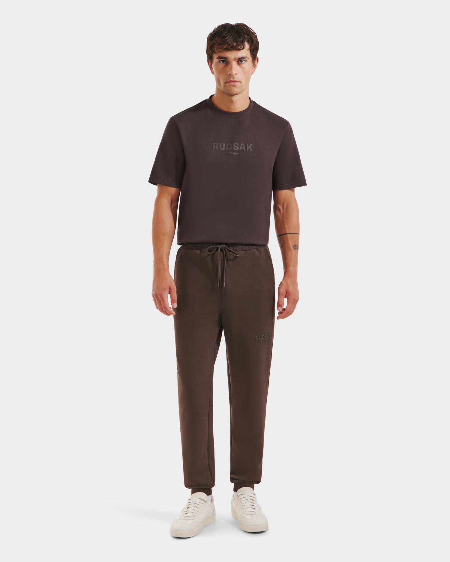 A man faces forward against a white background wearing Rudsaks JOSS - 9524009 Espresso M joggers with a matching dark brown short-sleeve tee and white sneakers.