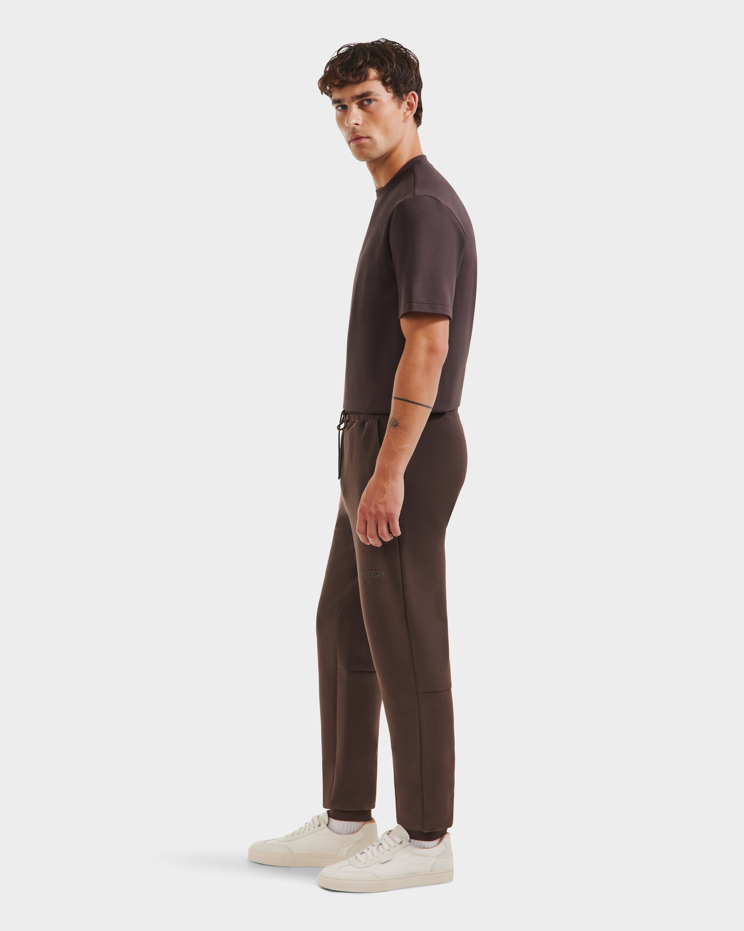 A man stands in profile wearing Rudsak’s JOSS - 9524009 Espresso M short-sleeve shirt and matching premium cotton joggers with white sneakers against a plain white background.