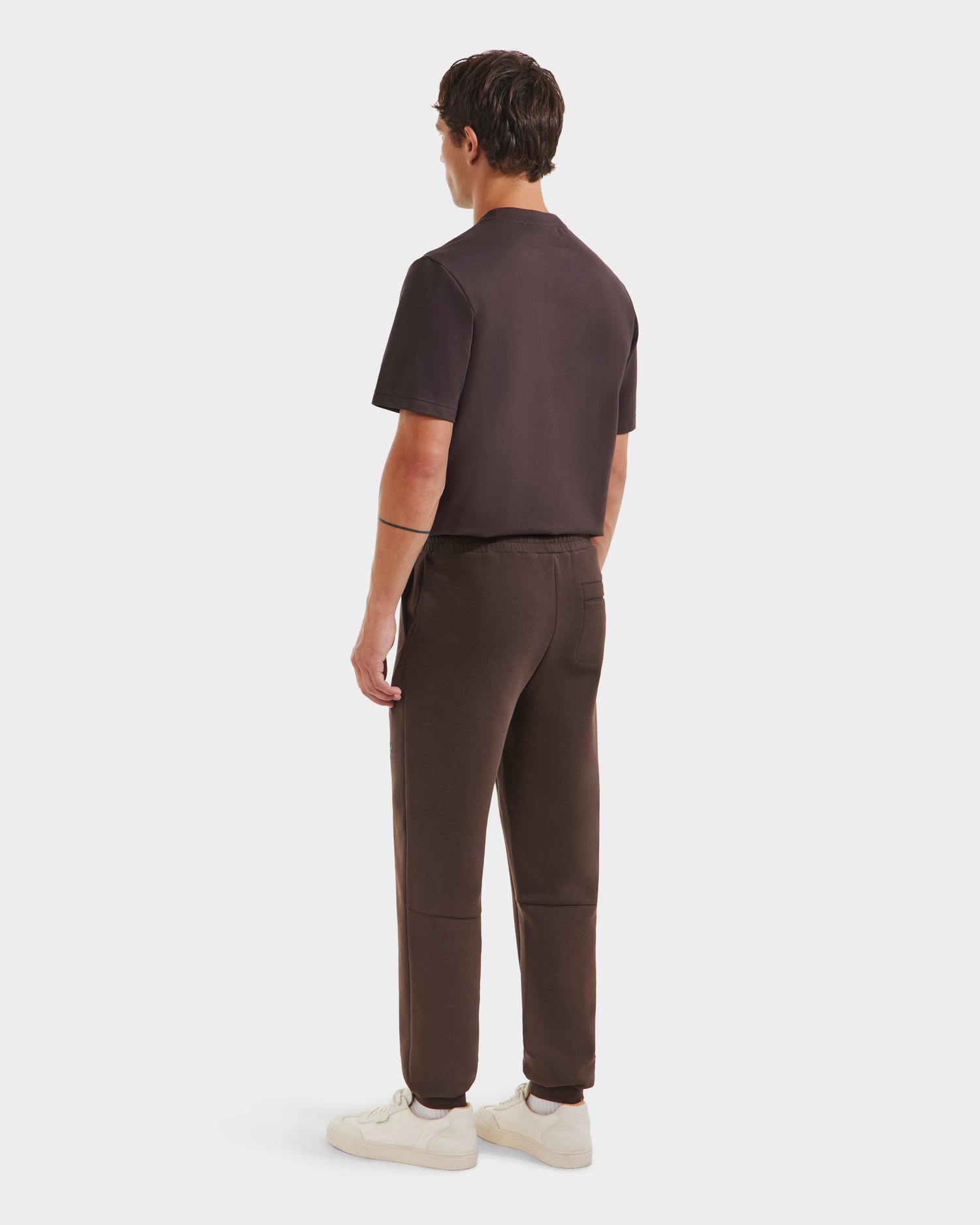 A person stands facing away from the camera, wearing the Rudsak JOSS - 9524009 Espresso M short-sleeve shirt and matching jogger pants, paired with white sneakers, against a plain white background.
