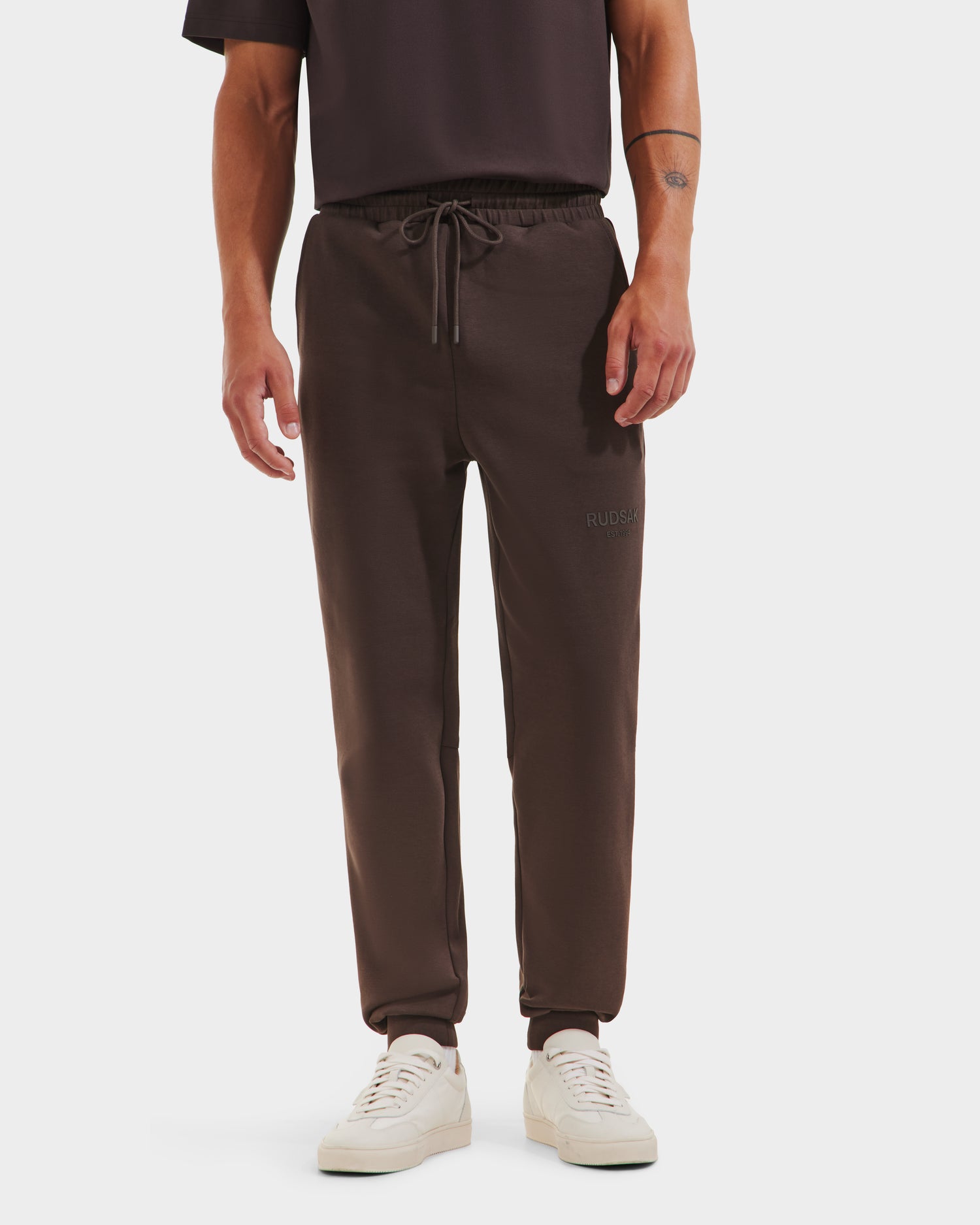 A person wears Rudsaks JOSS - 9524009 Espresso M unisex jogger pants with an elastic drawstring waistband, paired with a black T-shirt and white sneakers, standing against a plain white background.