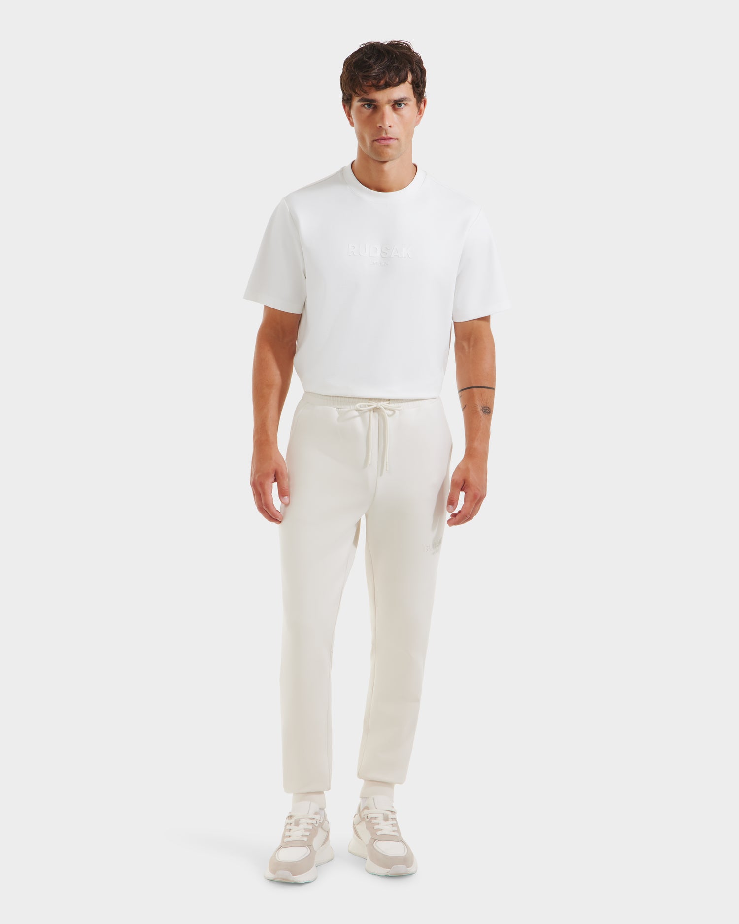 A man faces forward against a plain light background, wearing the Rudsak JOSS - 9524009 Vanilla M premium cotton joggers in cream, a white short-sleeve t-shirt, and beige-white sneakers.