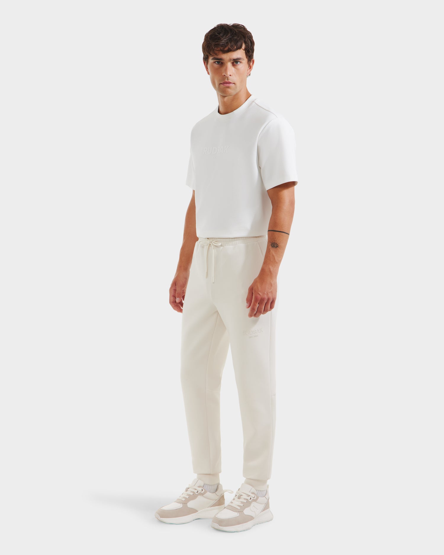 A man stands slightly angled, wearing Rudsaks JOSS - 9524009 Vanilla M t-shirt and premium cotton joggers with beige-and-white sneakers, set against a plain white background.