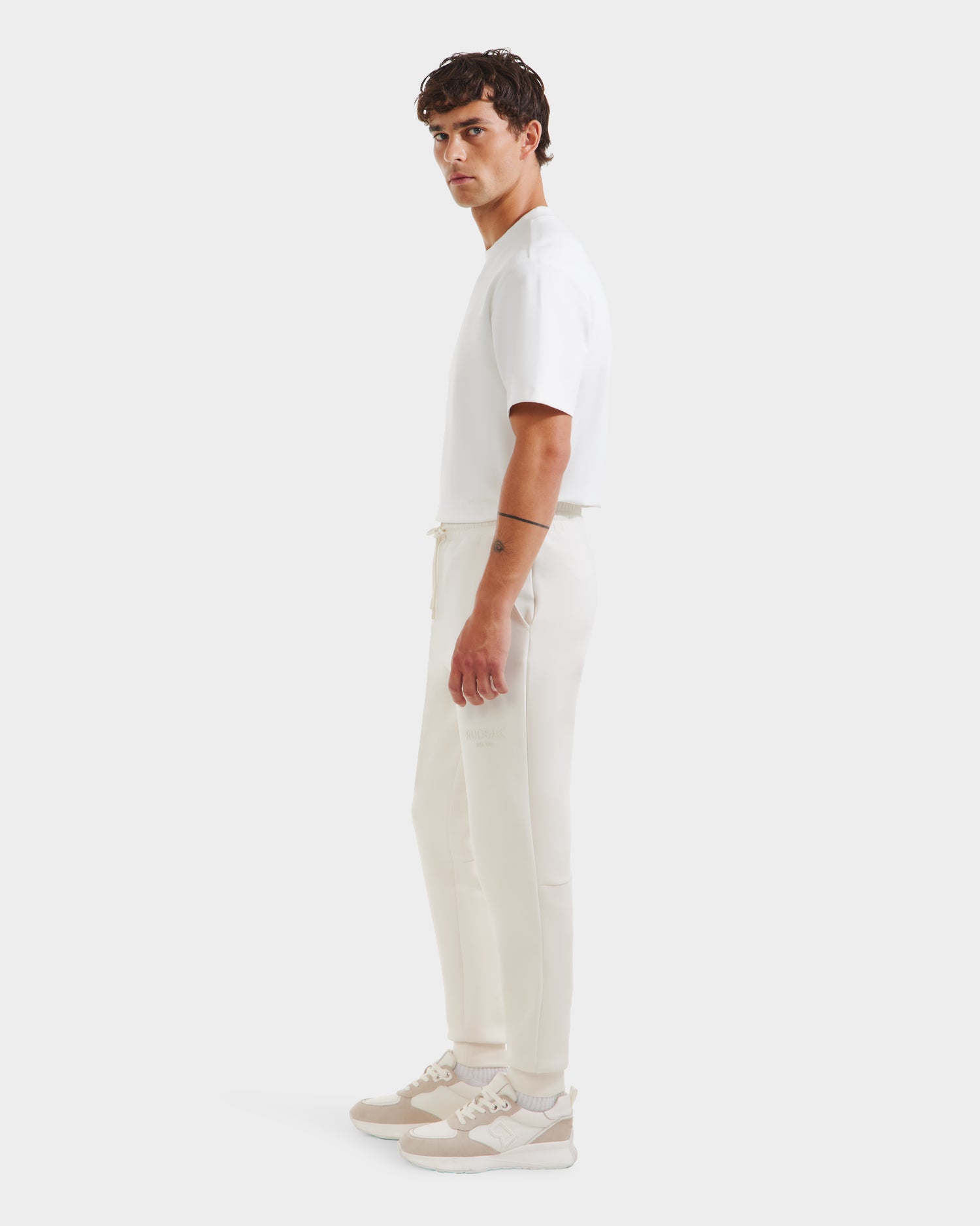 A man with short, dark hair and a neutral expression stands in profile against a white background, wearing Rudsaks JOSS - 9524009 Vanilla M premium cotton joggers, a short-sleeve shirt, and beige-and-white sneakers.