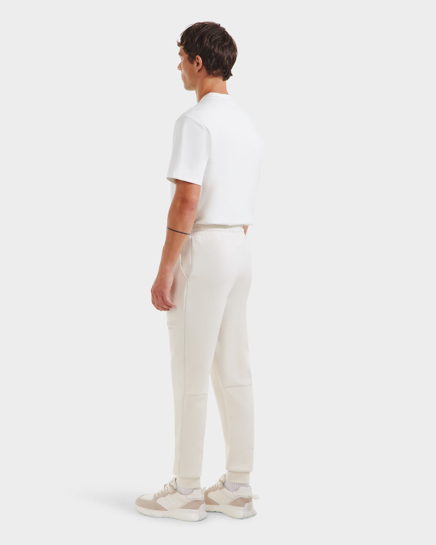 A person stands facing away in a white short-sleeve shirt, Rudsak JOSS - 9524009 Vanilla M premium cotton joggers, and white sneakers against a plain light background, looking to the side.
