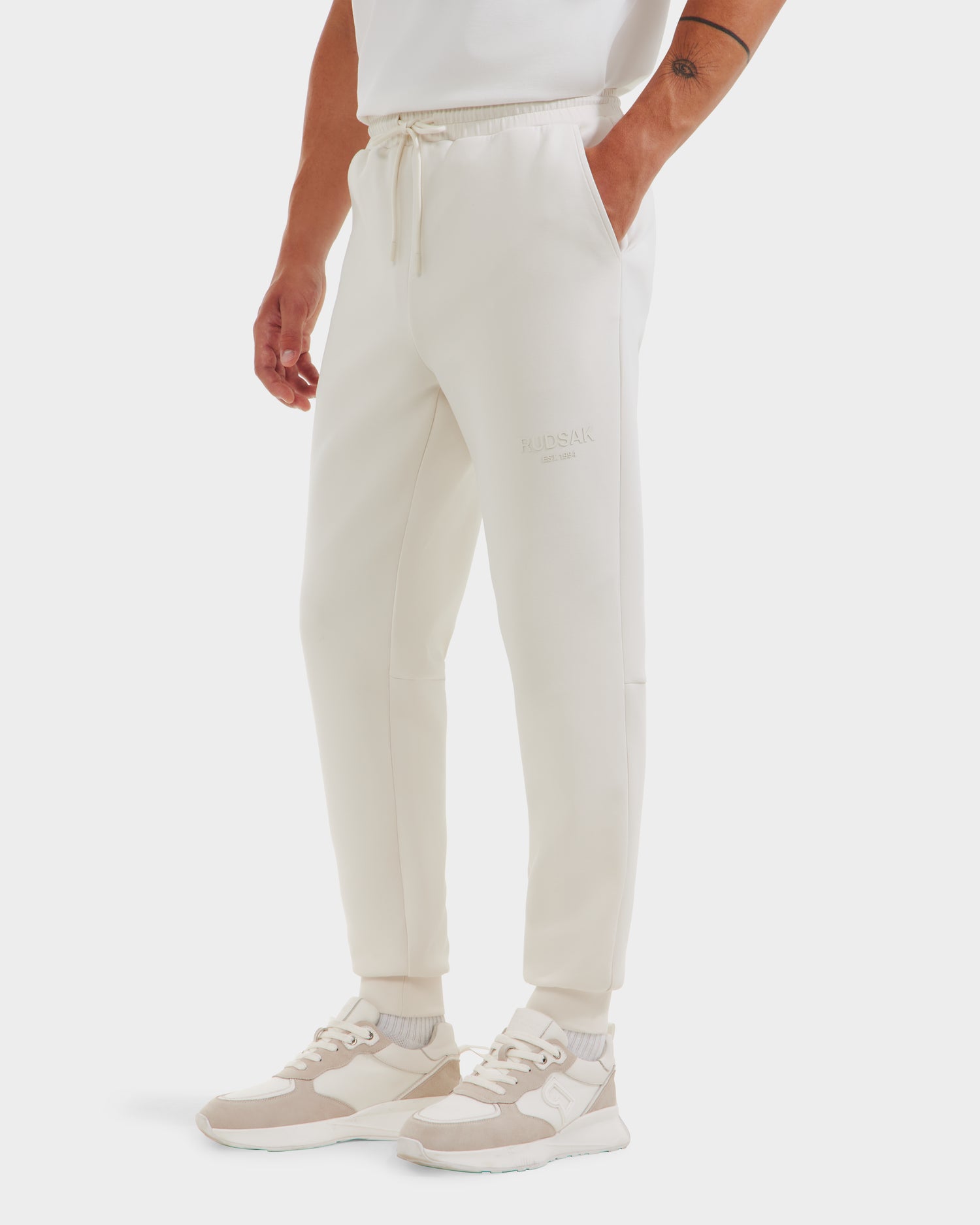 A person wears Rudsak JOSS - 9524009 Vanilla M jogger pants with a drawstring waist and side pockets, paired with white socks, white-beige sneakers, and a partially visible white shirt. Hands are in the joggers pockets.