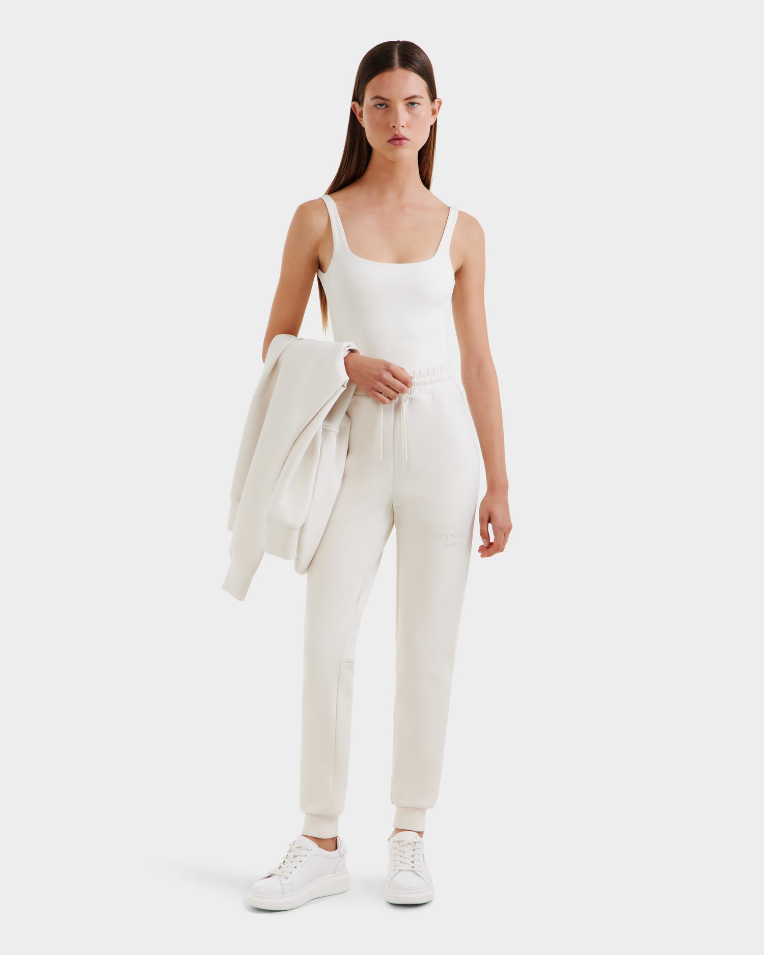 A woman with straight brown hair wears the Rudsak JOSS - 9524009 Vanilla W premium cotton jogger pants and a white sleeveless top, holding a white jacket over her arm while standing against a plain white background.