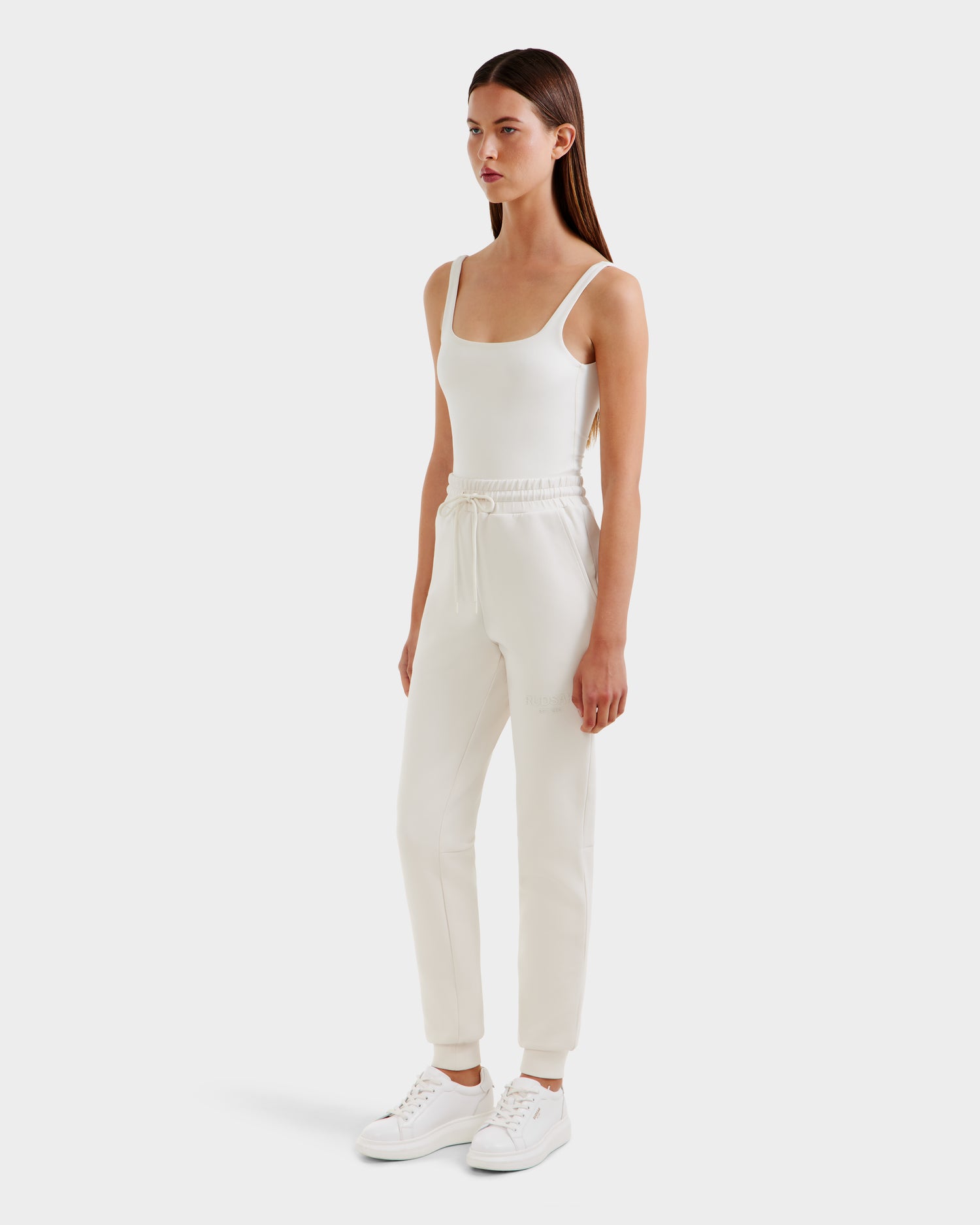 A woman with straight brown hair and a neutral expression wears the Rudsak JOSS - 9524009 Vanilla W fitted white tank top and high-waisted cotton joggers, paired with white sneakers, against a white background.