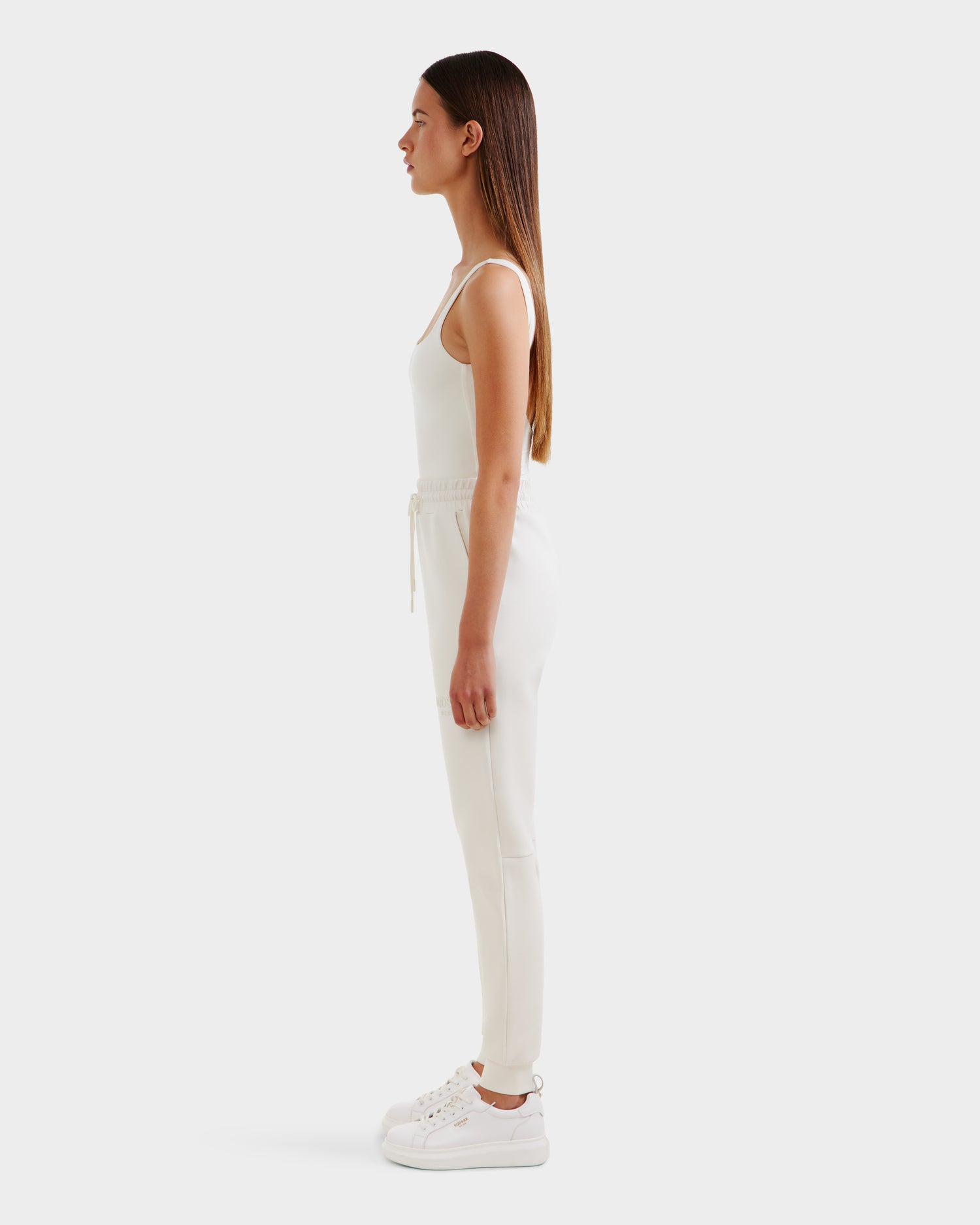 A woman with straight brown hair stands in profile, wearing Rudsak’s JOSS - 9524009 Vanilla W sleeveless top, premium cotton jogger pants, and white sneakers against a plain white background.