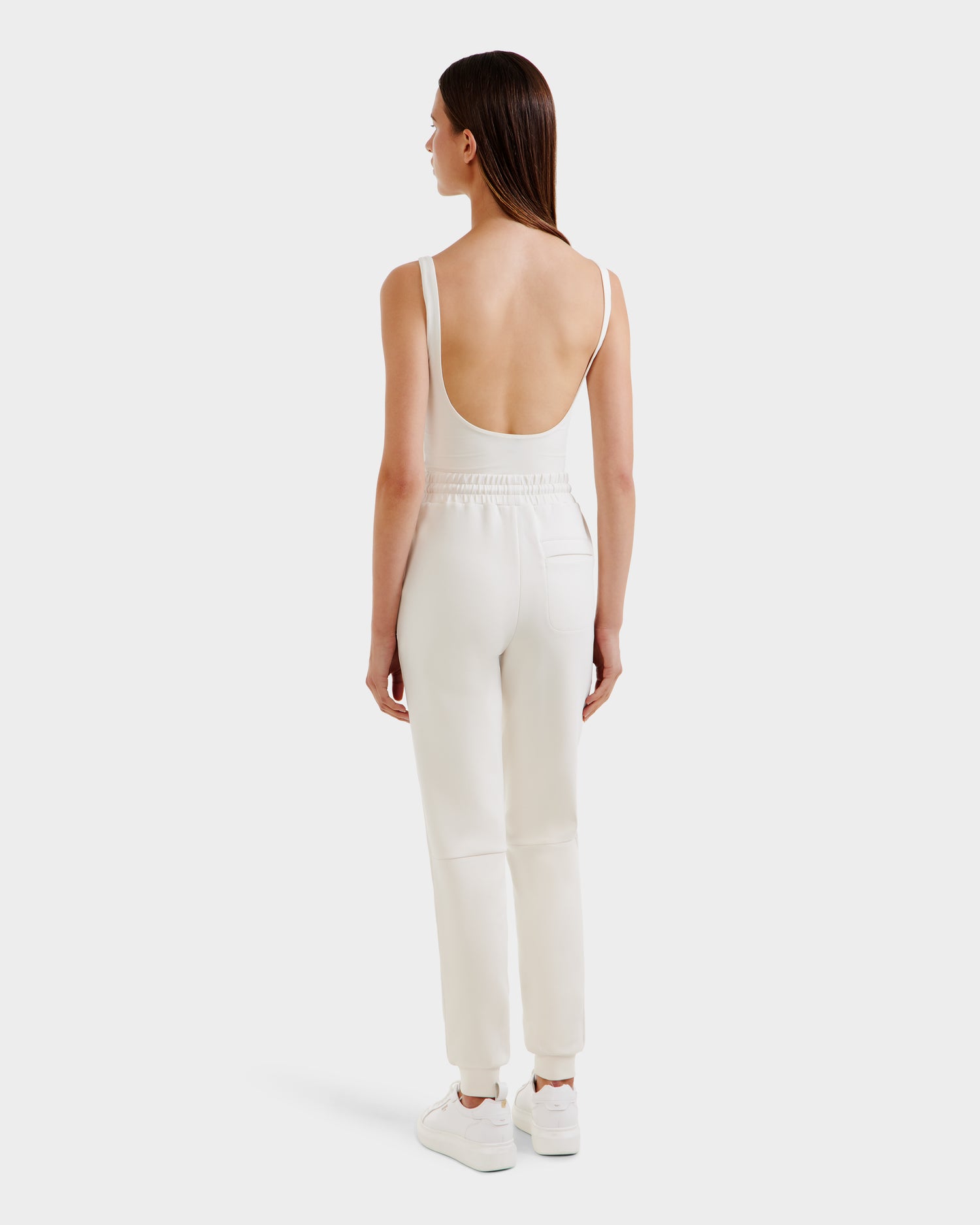 A woman with straight brown hair, seen from behind, models the JOSS - 9524009 Vanilla W jogger pants by Rudsak with a white low-back tank top and white sneakers against a plain light gray background.