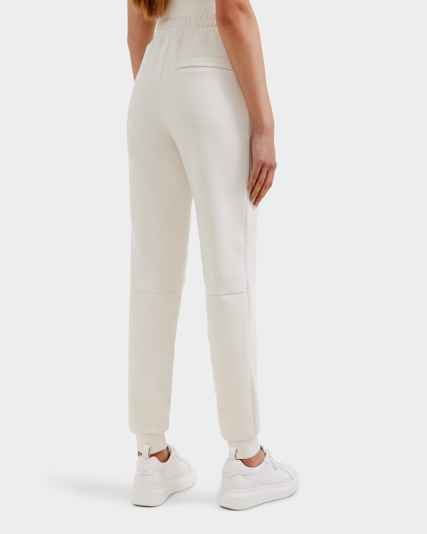 A person stands facing away, wearing Rudsaks JOSS - 9524009 Vanilla W unisex jogger pants and white sneakers. The premium cotton joggers have an elastic waistband, back pocket, and cuffed ankles. The backdrop is plain white.