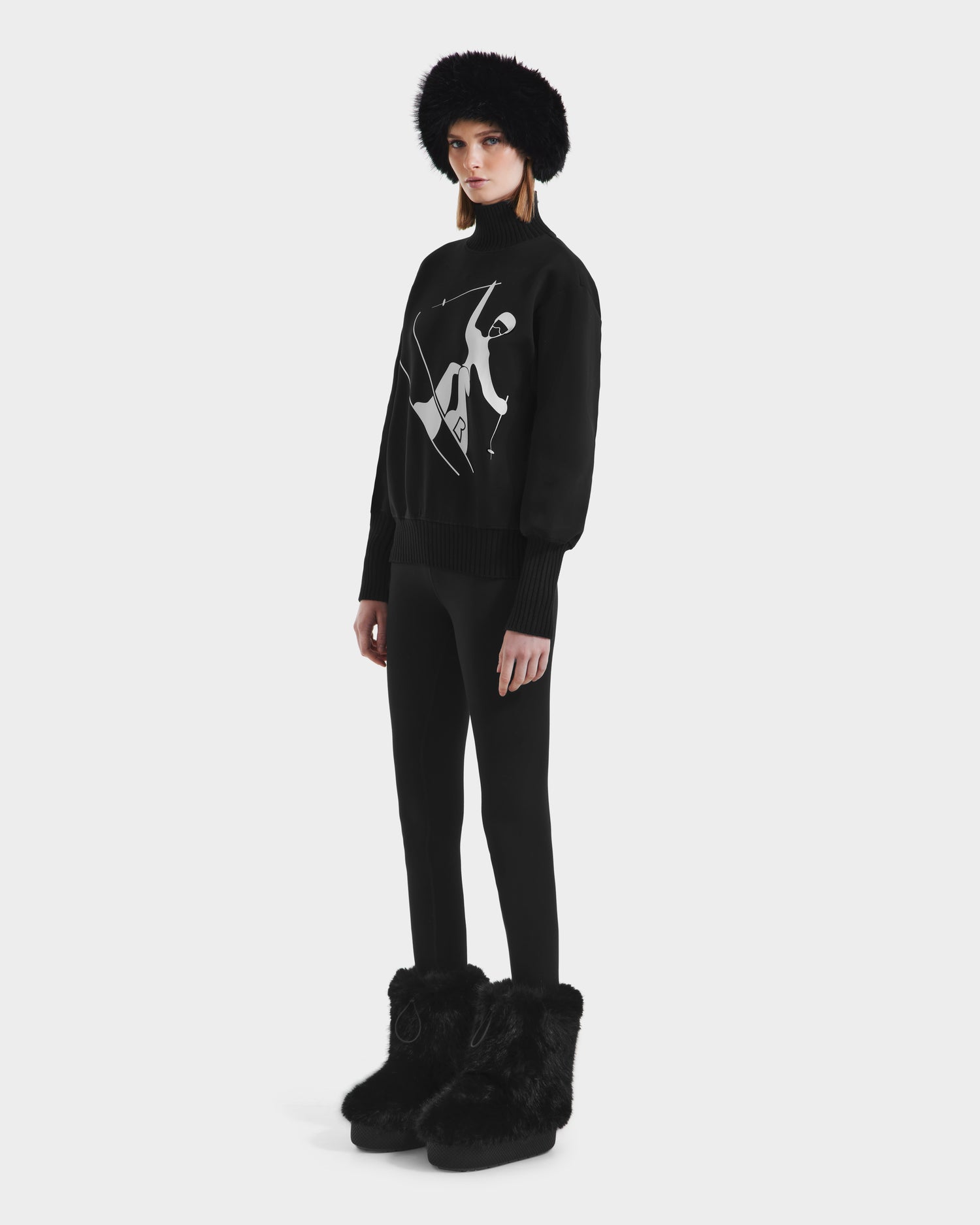 Against a plain white background, a person wears the Rudsak JOYCE - 8525647 Black sweater with an abstract white design, paired with black leggings, furry boots, and a black furry hat for a modern monochromatic look.
