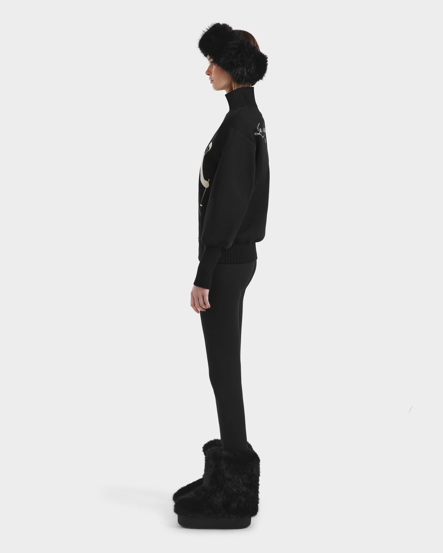 Wearing the Rudsak JOYCE - 8525647 Black après-ski sweater with a moon motif, fitted pants, black furry boots, and a matching black fur headband, a person stands in profile against a plain white background.