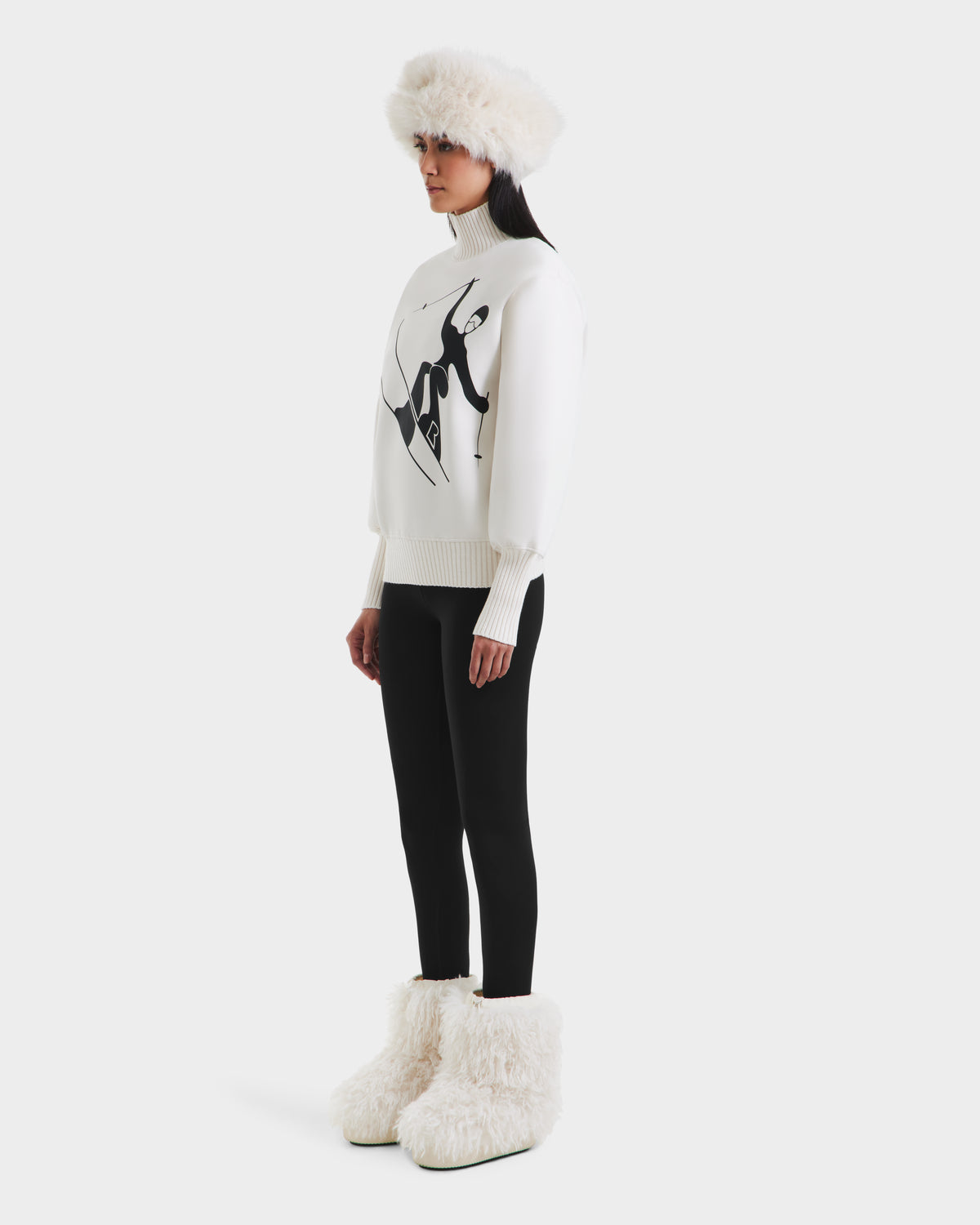 A woman stands sideways wearing the Rudsak JOYCE - 8525647 Vanilla women’s ski sweater, featuring a black abstract design, paired with black leggings, a white furry hat, and large fluffy boots against a plain background.