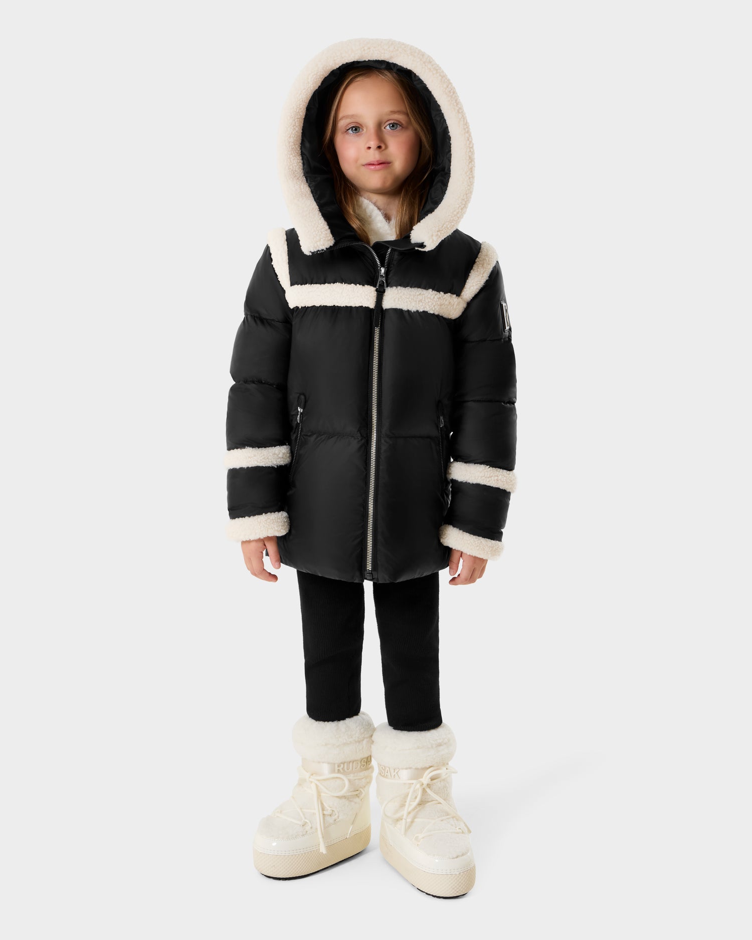 A young girl faces forward in a Rudsak JULIANA K - K125550 Black puffer coat with white fleece trim, black pants, and white winter boots. The hood is up and the backdrop is plain light gray.