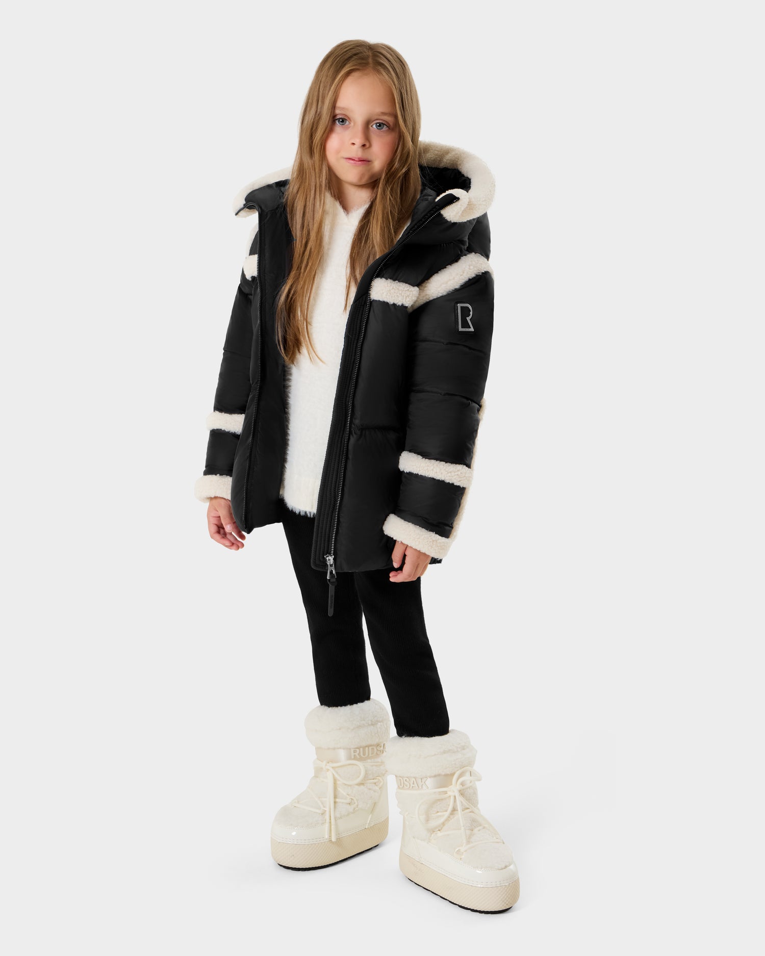A young girl with long light brown hair models the Rudsak JULIANA K - K125550 Black puffer jacket with white faux fur trim, paired with black leggings and white fluffy boots, standing against a plain light gray background.