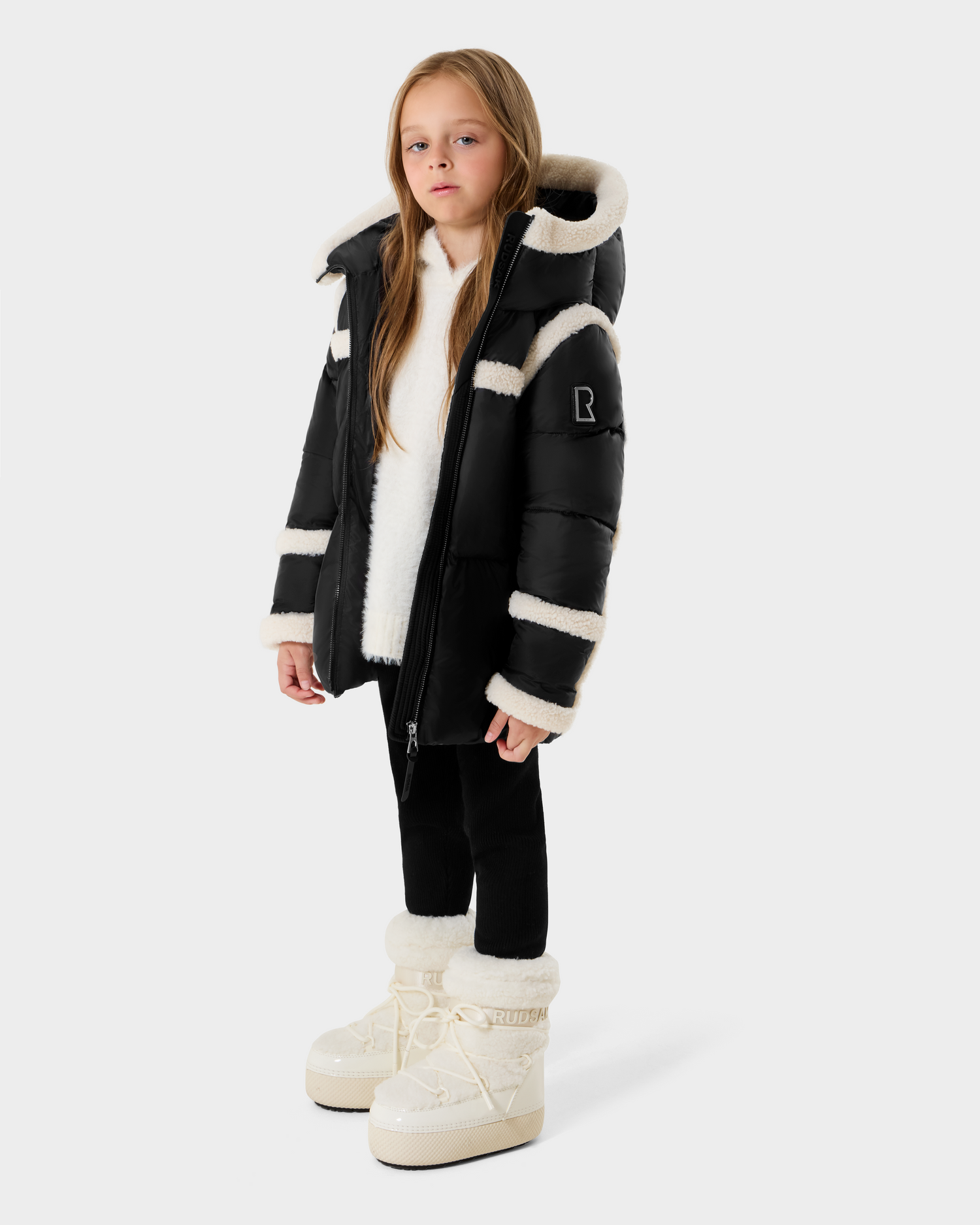 A young girl with long light brown hair and a neutral expression stands against a plain light background, wearing the Rudsak JULIANA K - K125550 Black puffer jacket, paired with black leggings and white fur-lined boots.