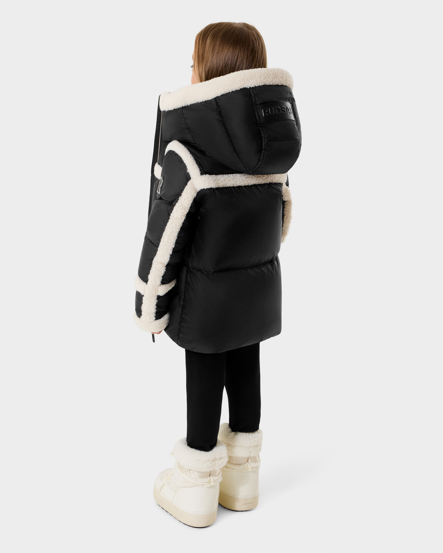 A young child models the Rudsak JULIANA K - K125550 Black puffer coat, featuring white shearling trim, paired with black leggings and white shearling boots, standing on a plain light background.