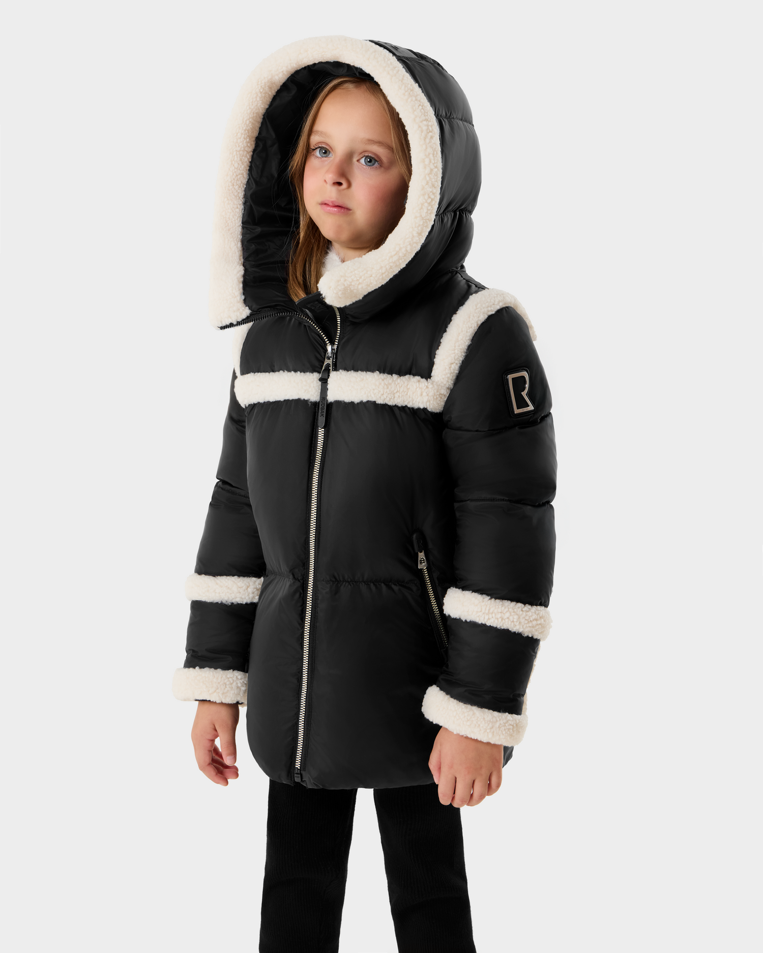 A young girl wears the JULIANA K - K125550 Black puffer coat by Rudsak, featuring white faux fur trim, a hood, vertical zipper, and an "R" patch on the sleeve against a light gray background.