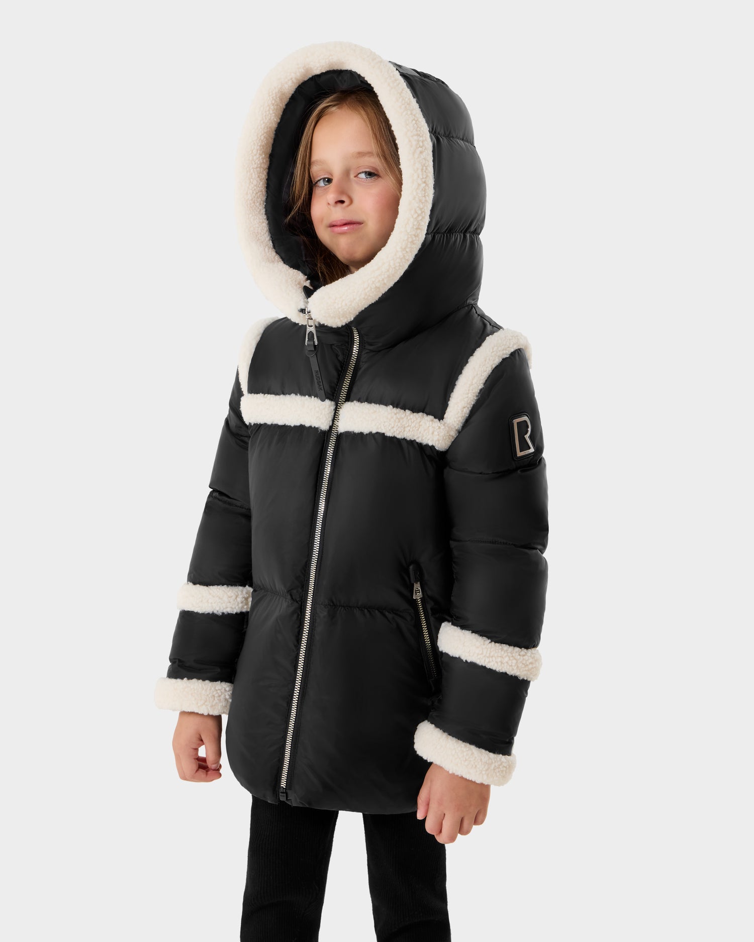 A young child wears the Rudsak JULIANA K - K125550 Black puffer jacket, featuring white faux fur trim on the hood, chest, sleeves, and cuffs, standing against a plain light gray background.