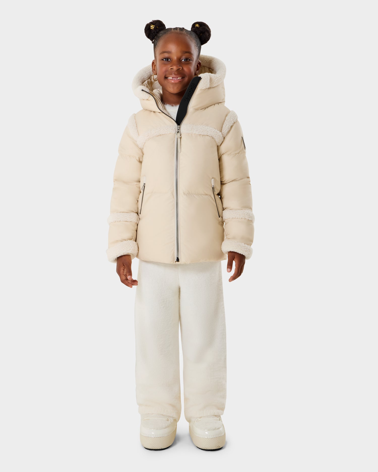 A young girl with braided hair styled in two buns smiles, wearing the Rudsak JULIANA K - K125550 Vanilla cream-colored puffer jacket with hood, matching wide-leg pants, and boots against a plain white background.