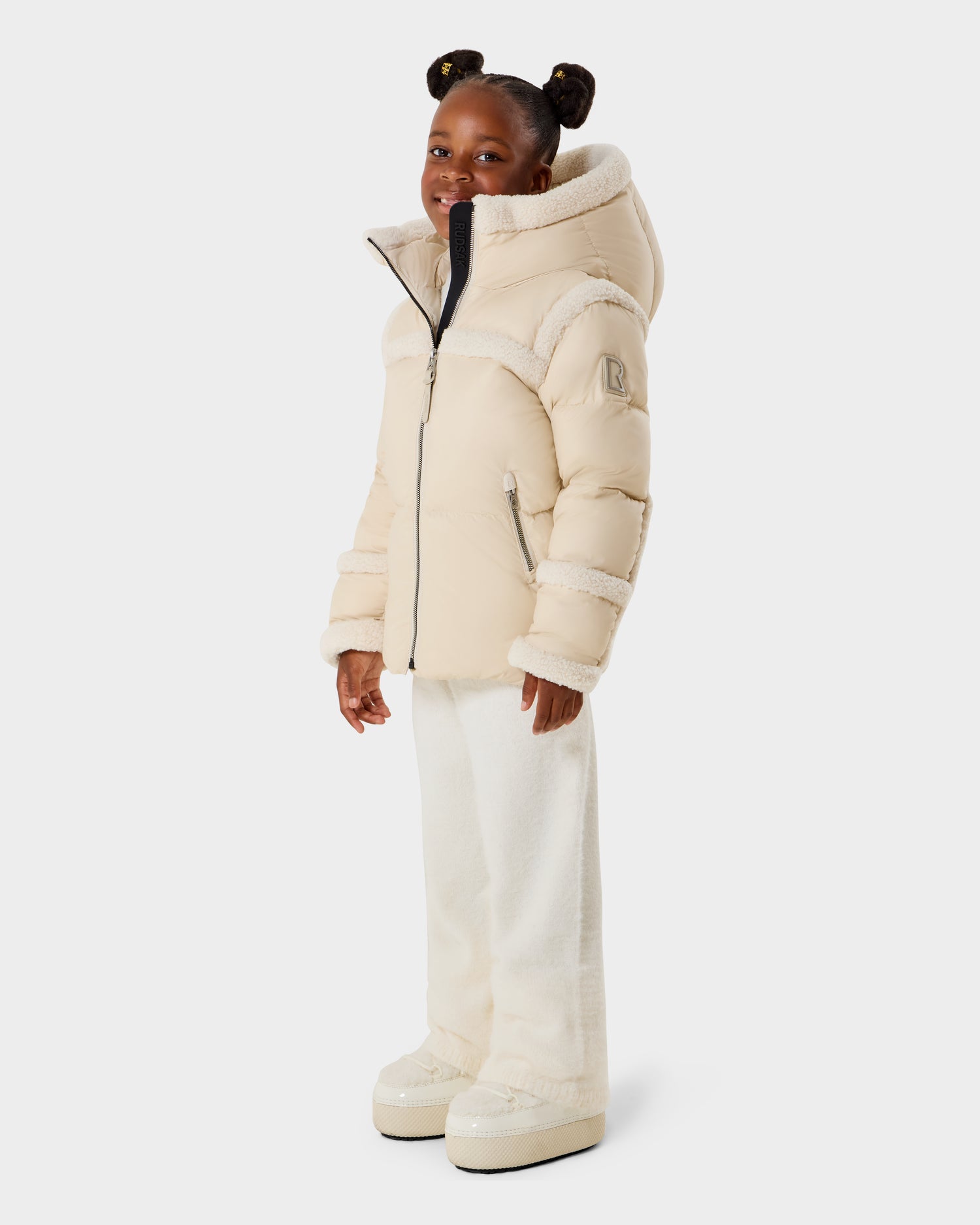 A young girl smiles in the Rudsak JULIANA K - K125550 Vanilla hooded puffer jacket, paired with white pants and beige platform shoes. Her hair is in two buns with bows, set against a plain white background.