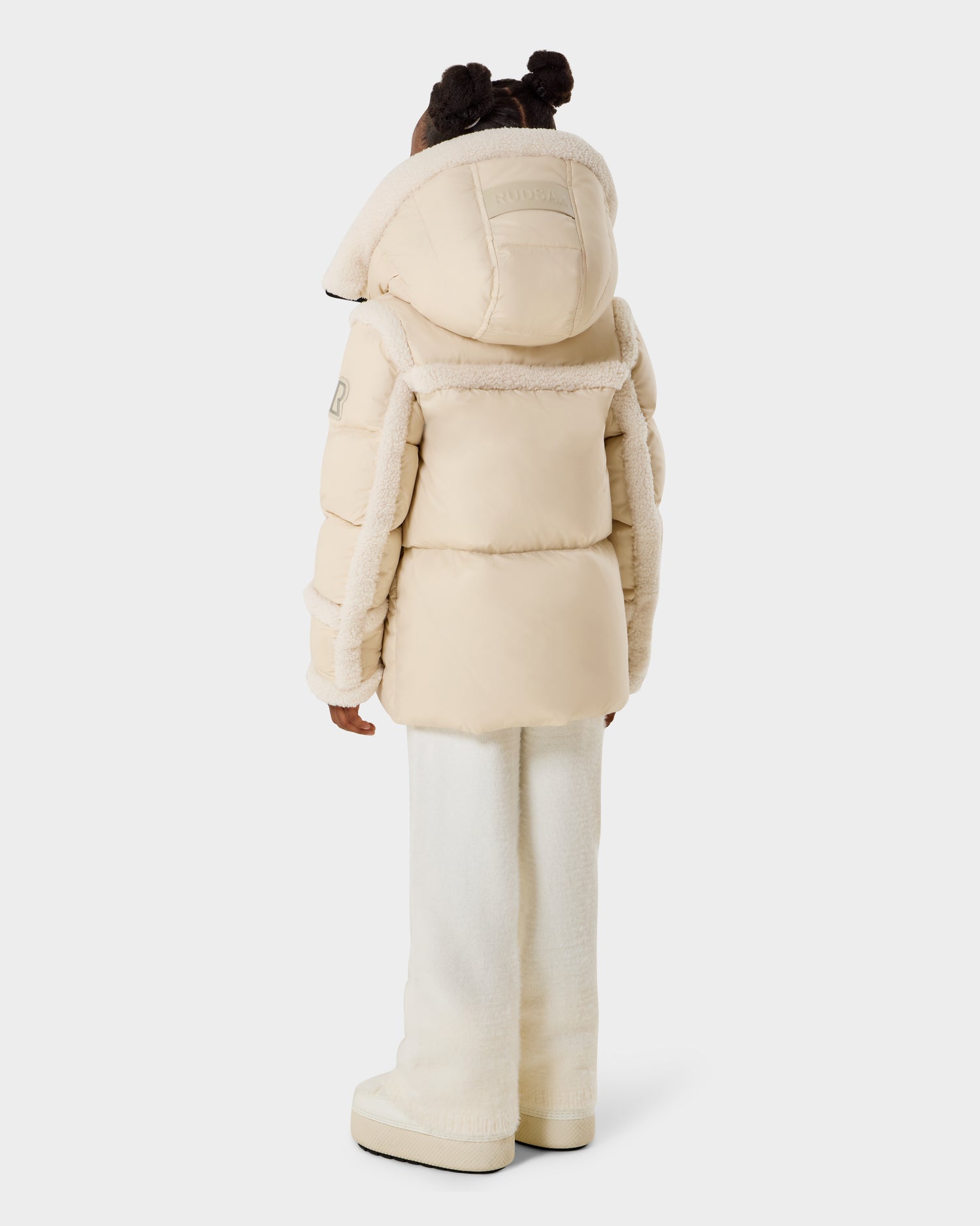 A child with their back to the camera wears the Rudsak JULIANA K - K125550 Vanilla cream-colored puffer jacket with a hood, styled with white pants and matching boots, against a plain light background.