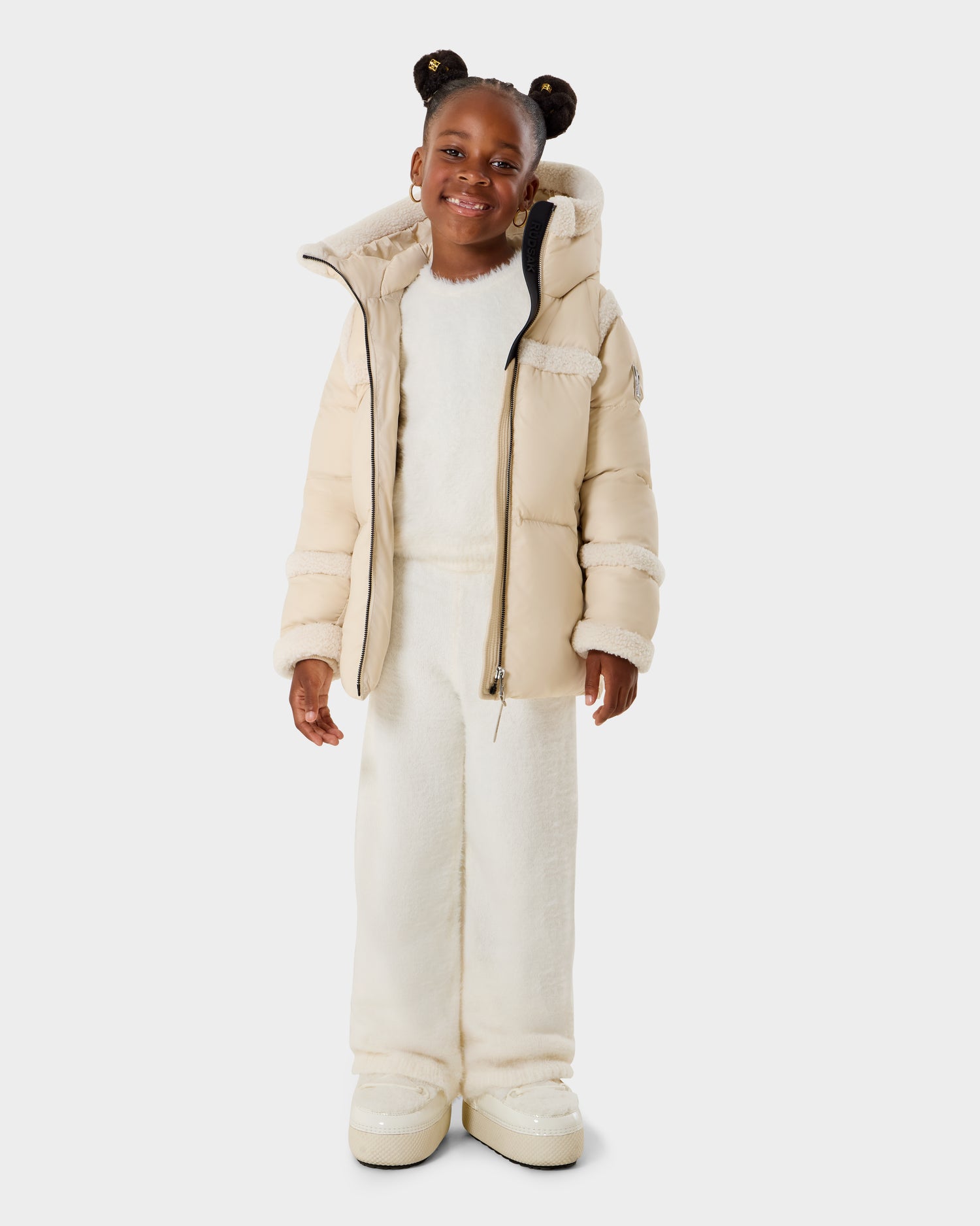 A smiling young girl with two hair buns wears the Rudsak JULIANA K - K125550 Vanilla puffer jacket over a white fuzzy outfit and matching white shoes, standing against a plain light background.