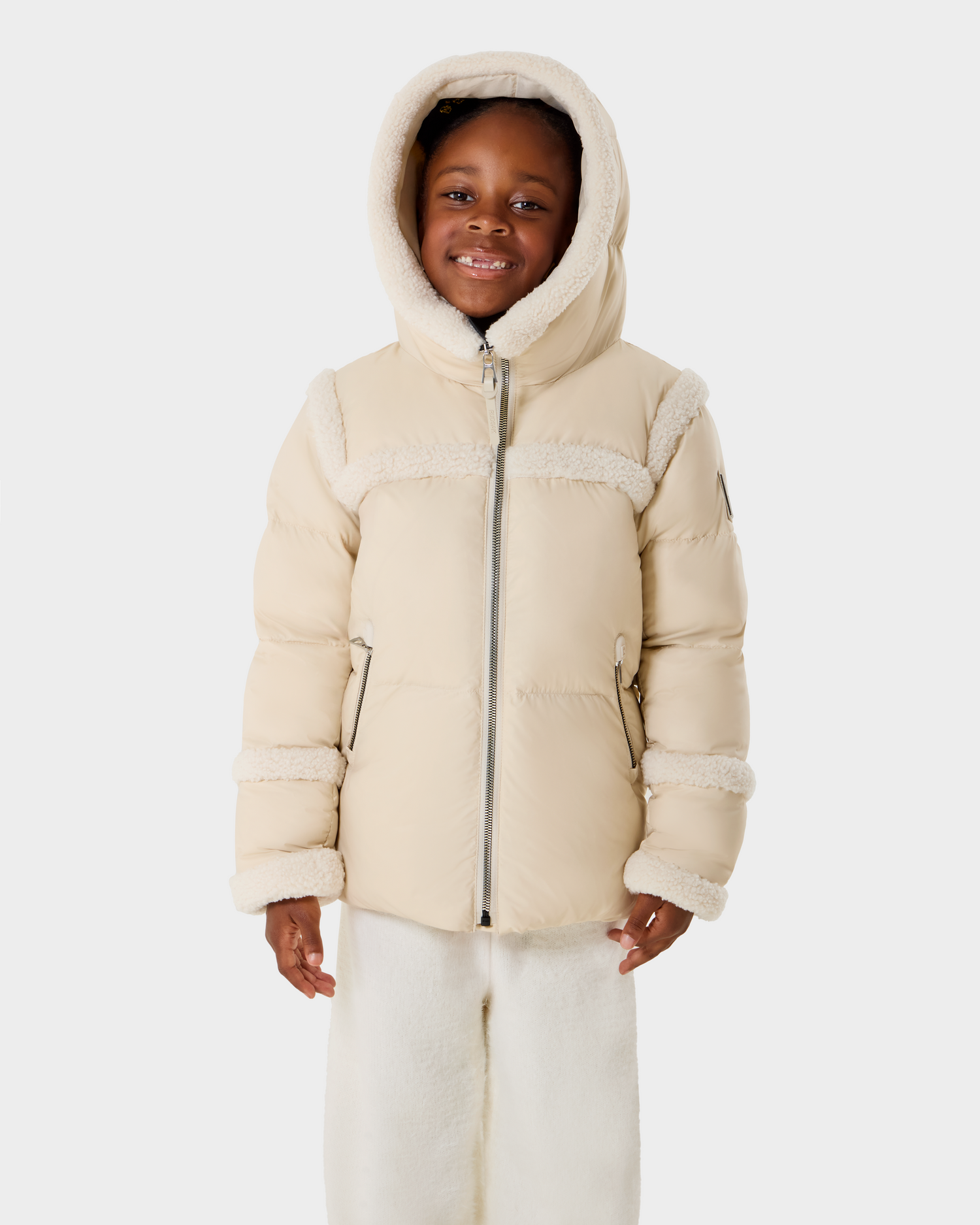 A young child wears the Rudsak JULIANA K - K125550 Vanilla, a light beige hooded winter jacket with fleece trim and matching cozy pants, smiling against a plain white background.