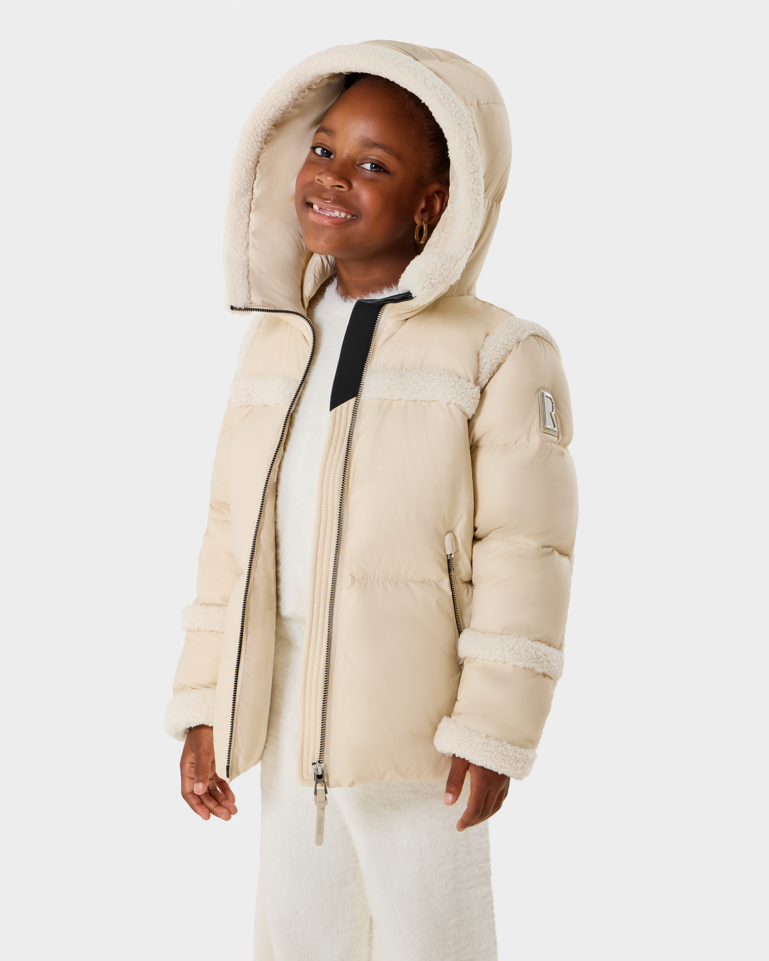 A young girl smiles in the Rudsak JULIANA K - K125550 Vanilla beige hooded puffer jacket with fleece details and a sleeve patch, partially unzipped over a white outfit against a plain white background.