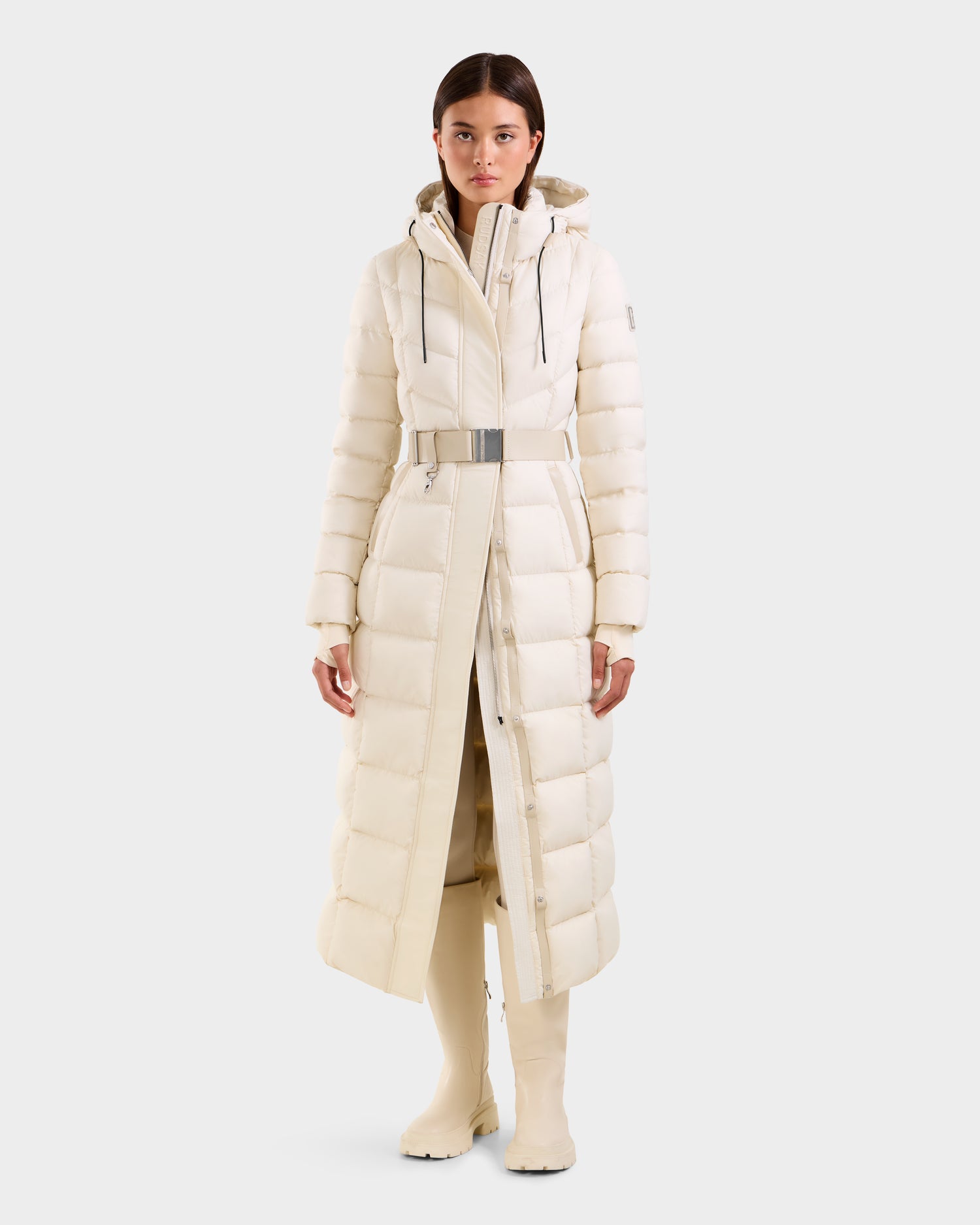 A woman faces forward, wearing the Rudsak JULIETA M-L - L125559 Vanilla long white maxi down puffer with a hood and silver belt, paired with matching high boots. The background is plain light gray.