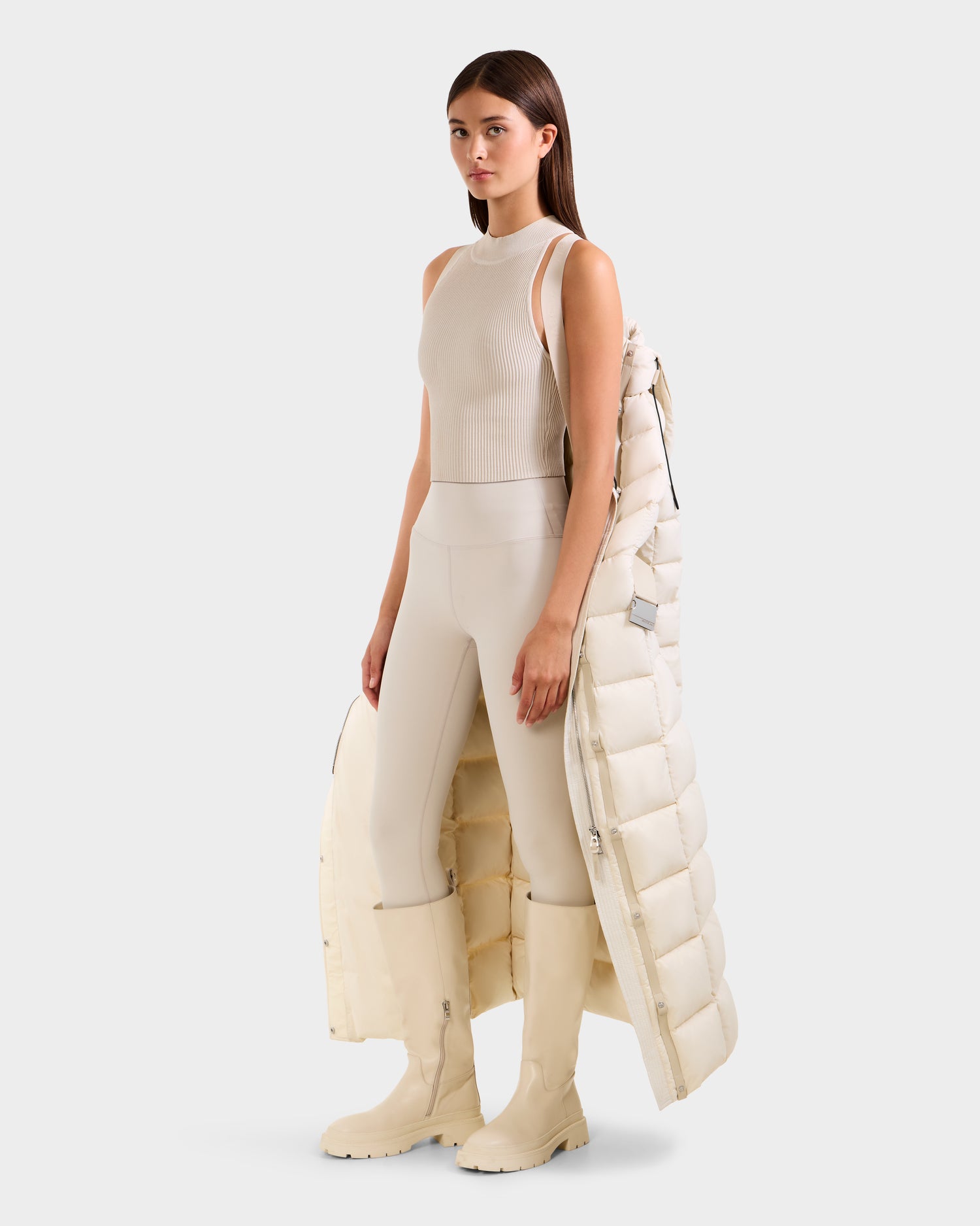 A woman stands against a plain light background, wearing neutral tones and holding the Rudsak JULIETA M-L - L125559 Vanilla maxi down puffer draped over one arm as her ideal winter layer.