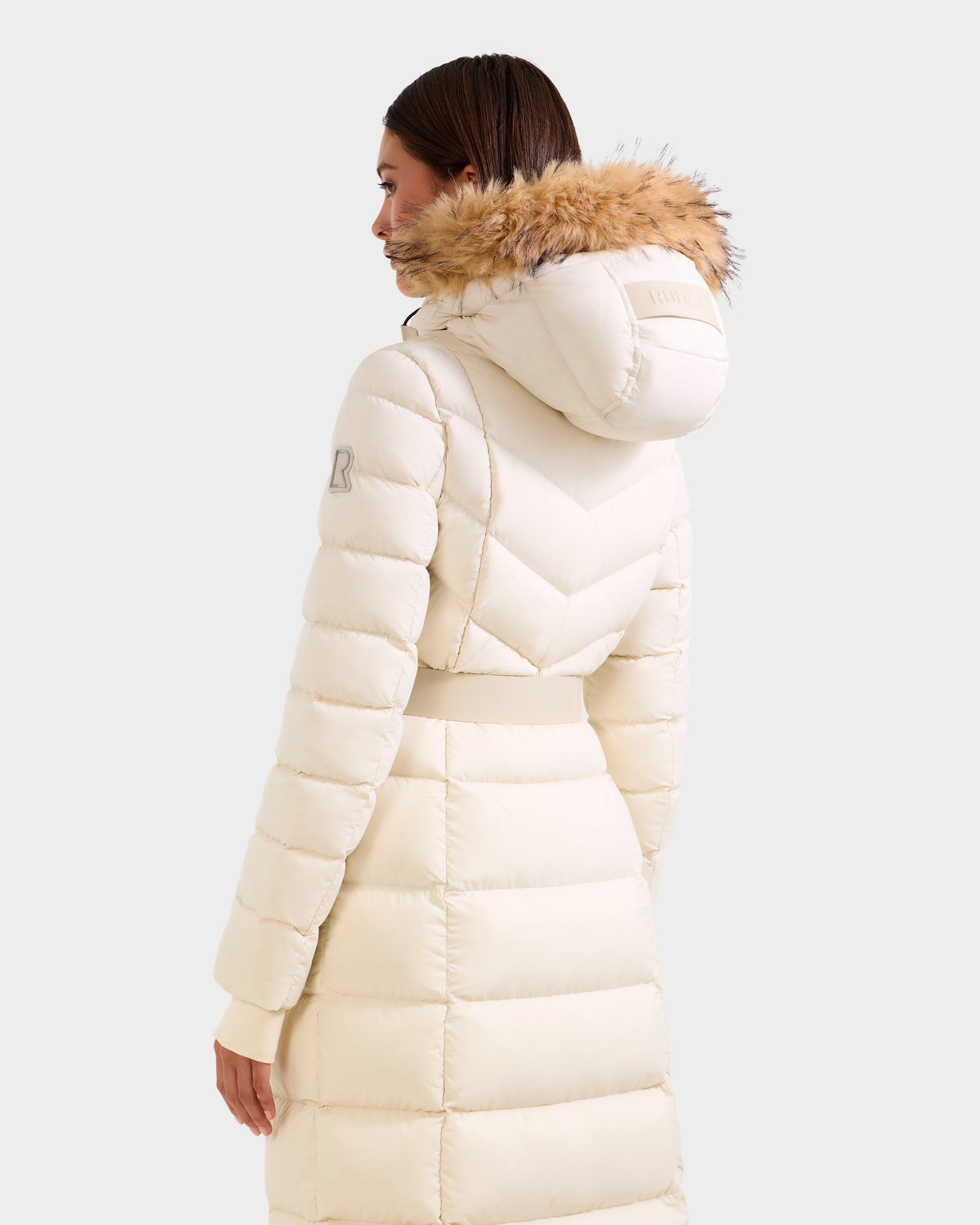 A woman with straight brown hair wears the Rudsak JULIETA M-L - L125559 maxi down puffer coat in vanilla, featuring a hood trimmed with light brown faux fur. She is turned slightly to show the back and side of this cozy winter layer.