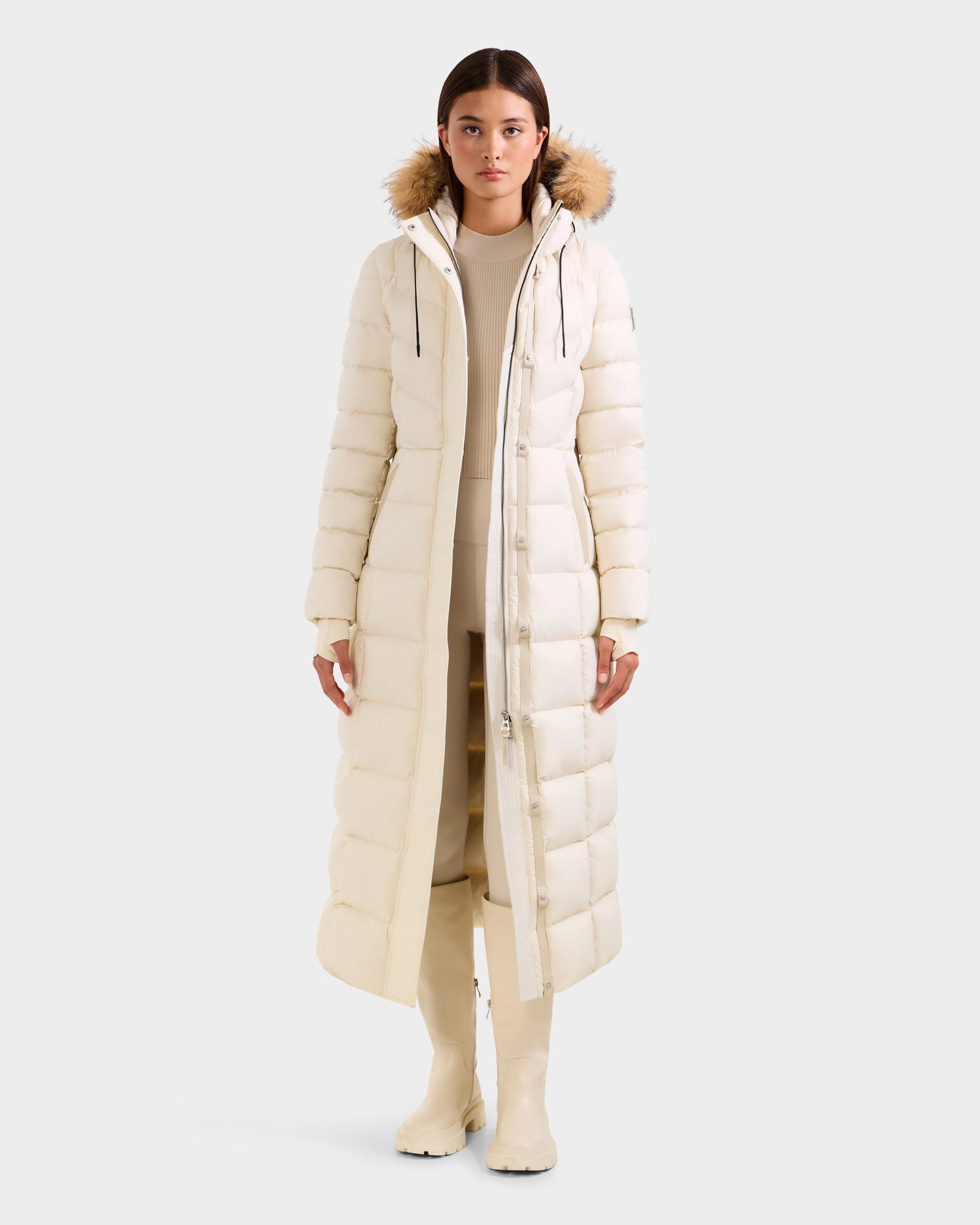 A woman faces forward wearing the Rudsak JULIETA M-L - L125559 Vanilla maxi down puffer with a fur-trimmed hood, paired with light pants and neutral knee-high boots, creating a chic winter look against a plain white backdrop.