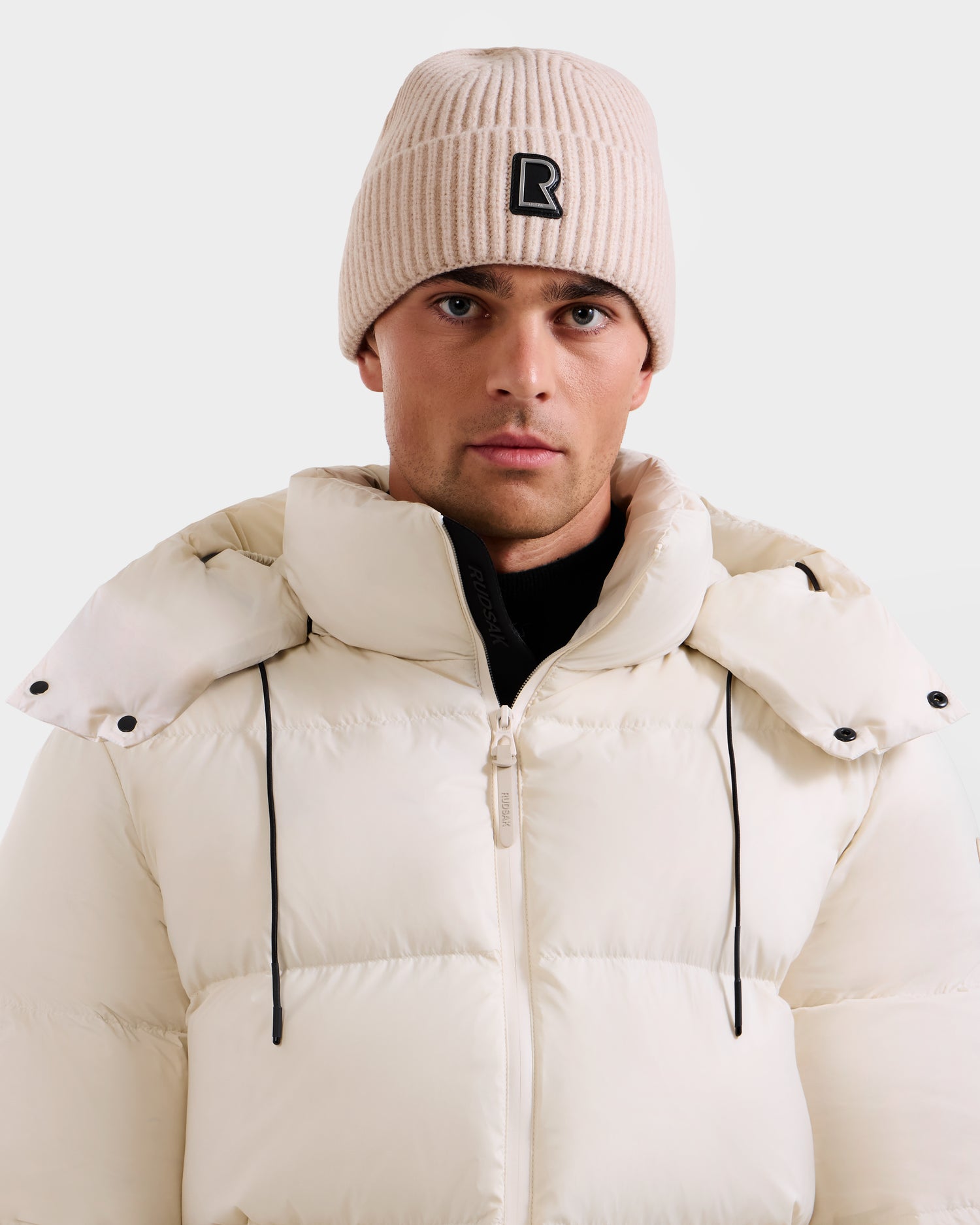 A man stands facing forward against a light background, wearing the Rudsak JULIO - U423502 Vanilla knit beanie and a white puffer jacket with black drawstrings, showcasing this stylish winter look.