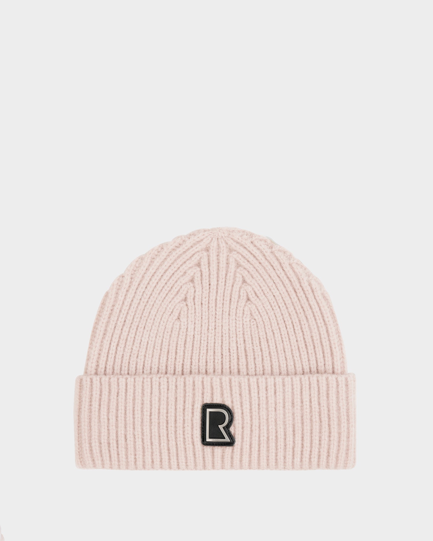 The JULIO - U423502 Vanilla by Rudsak is a light pink unisex knit beanie with a ribbed design, folded brim, and black-and-white letter R patch on the front, shown against a plain white background.
