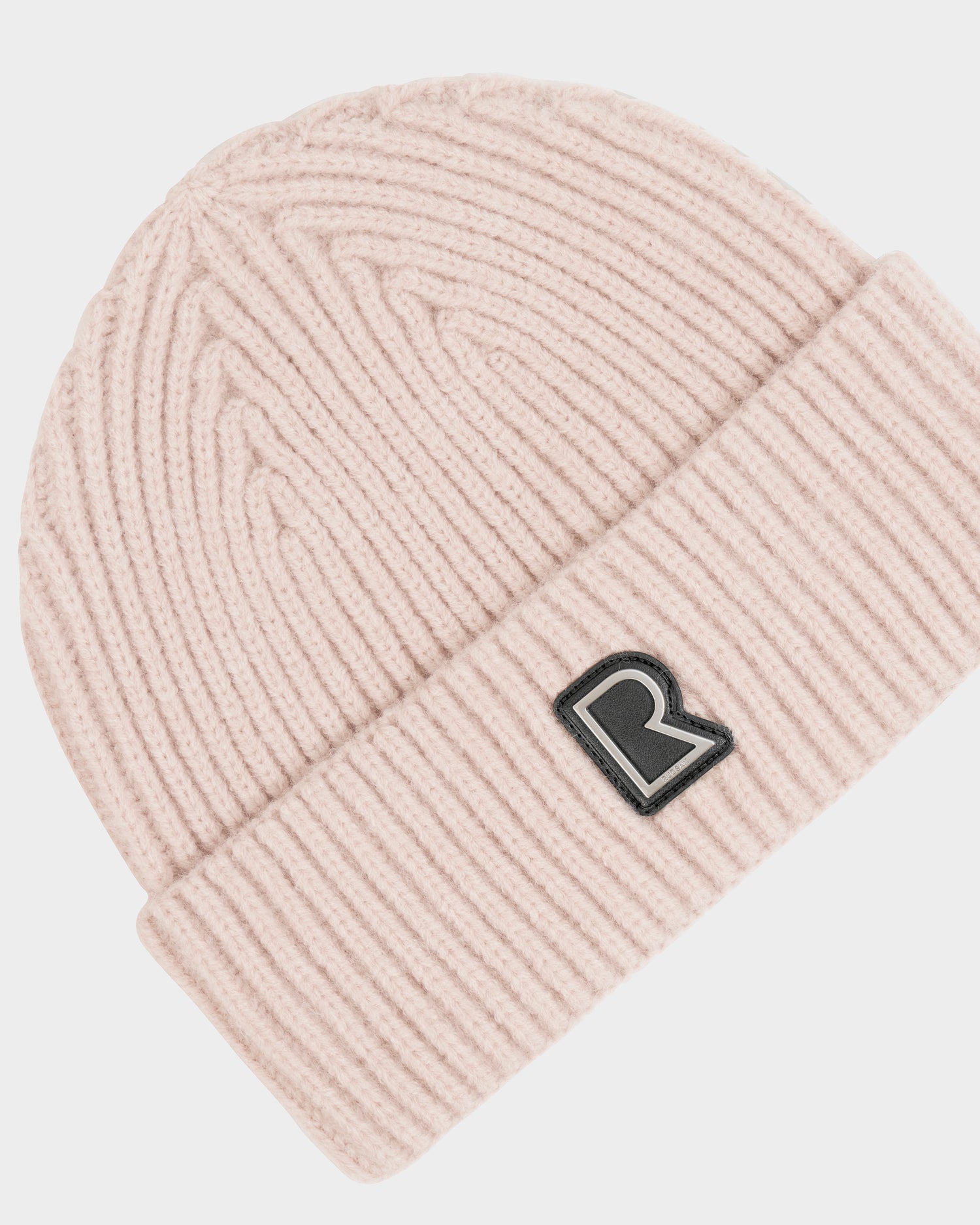 The JULIO - U423502 Vanilla by Rudsak is a light pink unisex knit beanie with a folded cuff and a black and white R patch on the front—a true winter essential.