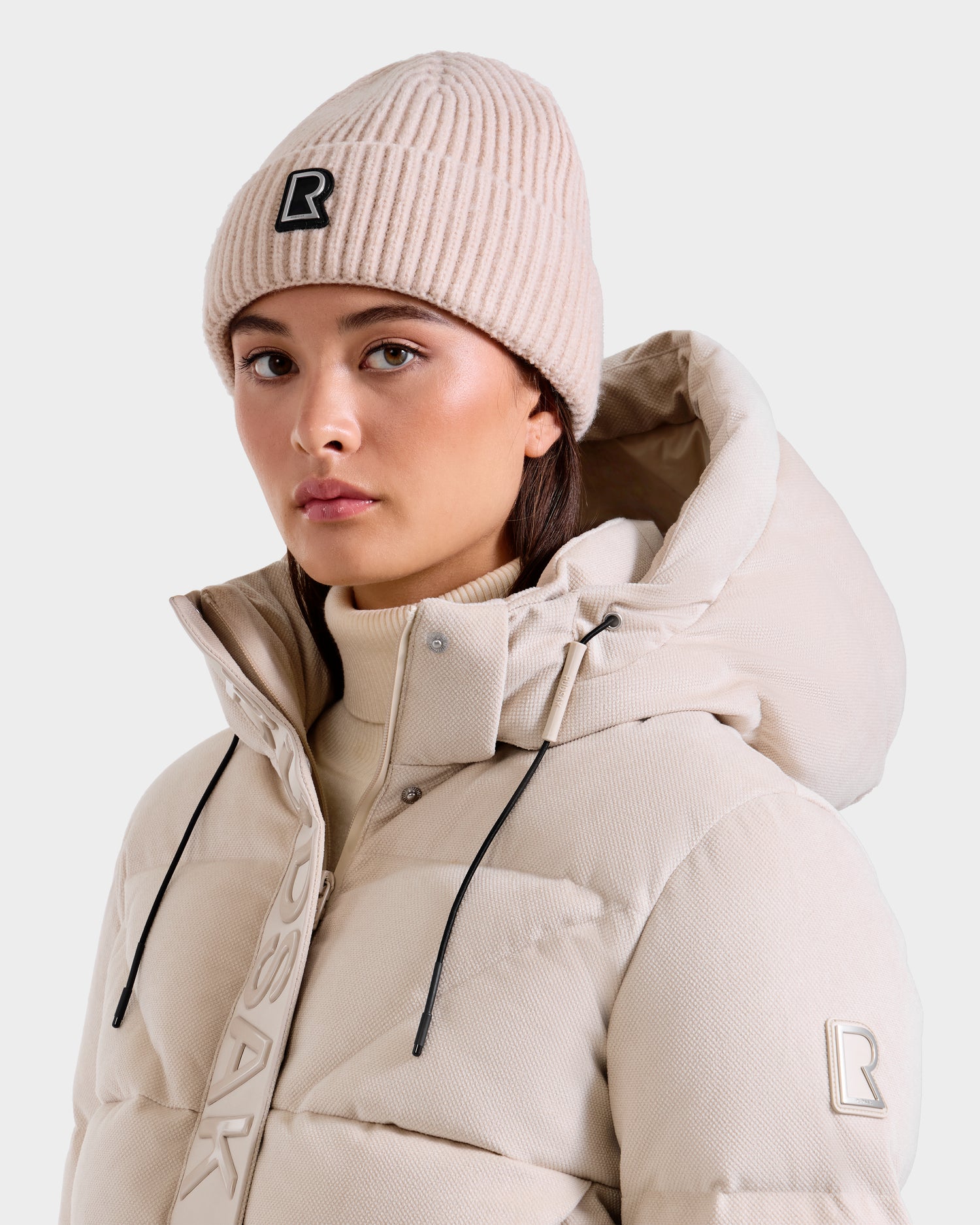 A woman wears the Rudsak JULIO - U423502 Vanilla puffer jacket with a high collar and hood, paired with a unisex knit beanie featuring a black and white “R” logo, standing against a plain background for a true winter essential look.