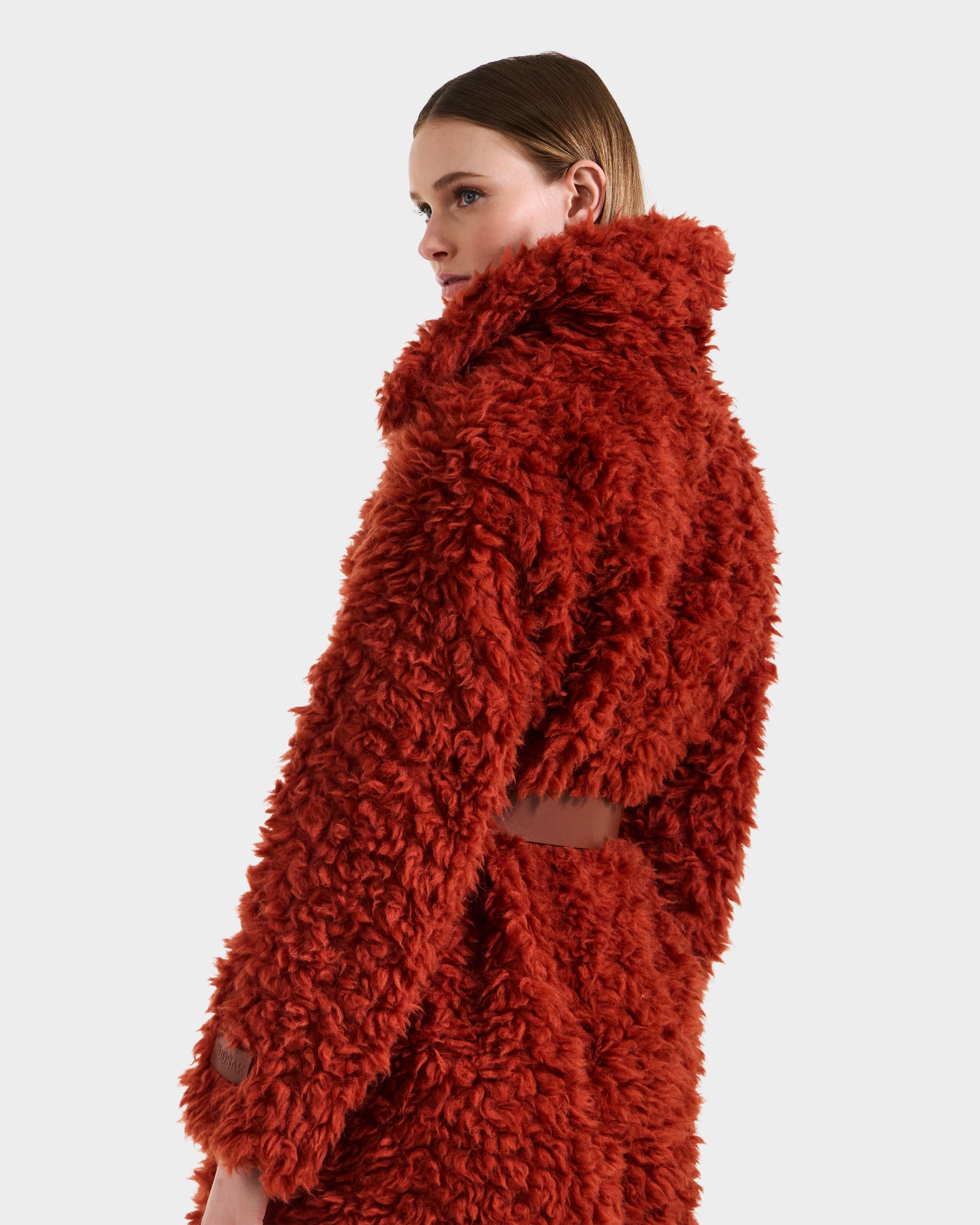 A person wearing the KAMILAH - 8125609 Rust trench by Rudsak, a textured women’s faux fur coat with a high collar and soft, curly look, gazes to the side against a plain light background.