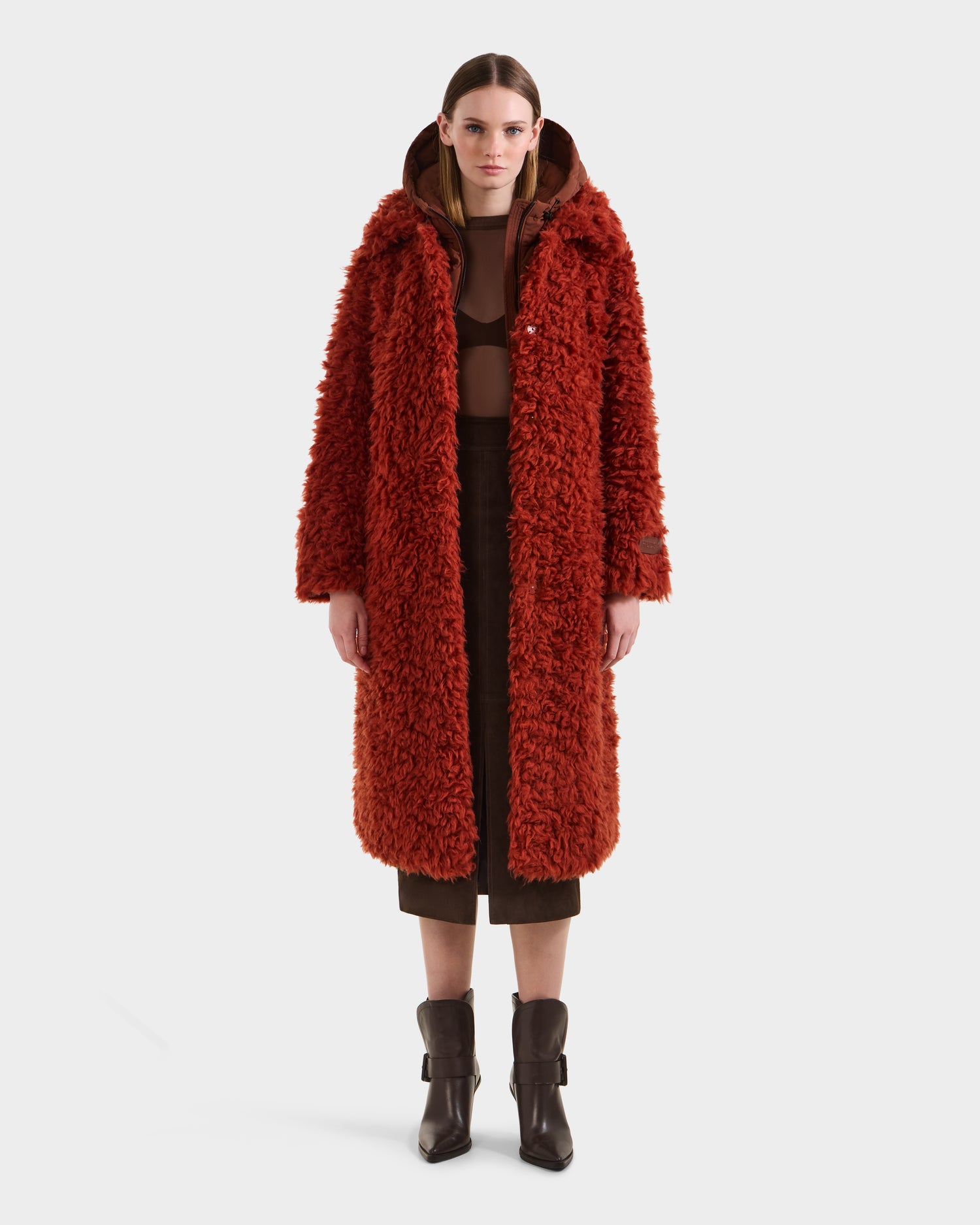 A woman stands against a white background wearing the Rudsak KAMILAH - 8125609 Rust long faux fur trench over a brown outfit with a hoodie and calf-high brown boots, facing forward with a neutral expression.